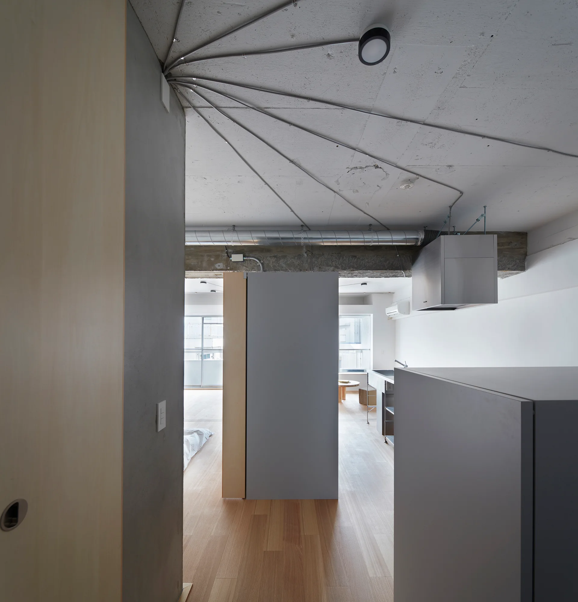 A minimalist apartment interior with light wood floors, grey divider cabinets, and radiating metal electrical conduits on a raw concrete ceiling.