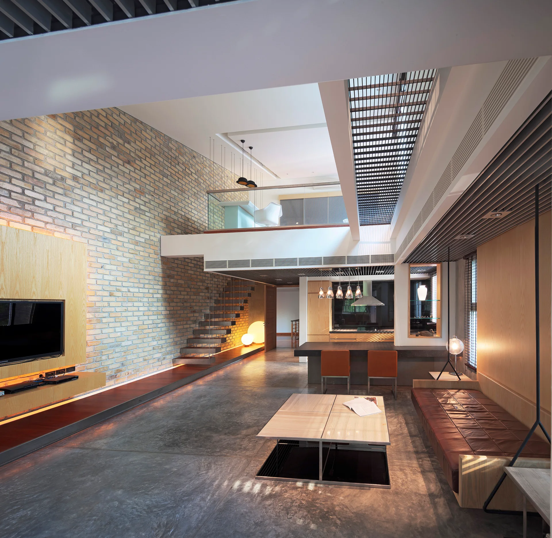A spacious multi-level living room features a large exposed brick wall, a floating wooden staircase, polished concrete flooring, and a mezzanine balcony.