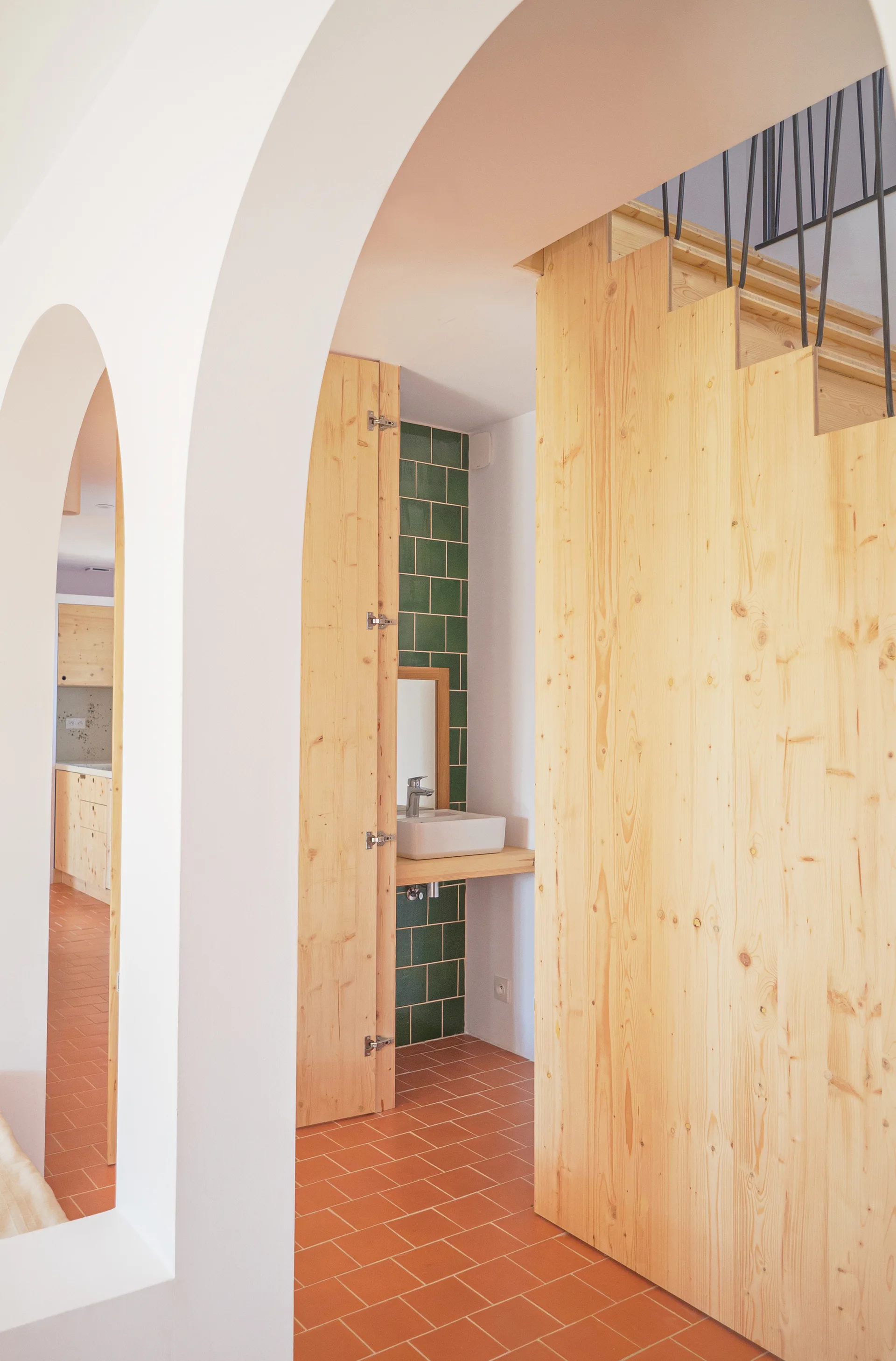 A white arched doorway reveals a hallway with terracotta tiles, light pine wood paneling, and a small vanity area with dark green wall tiles and a sink.