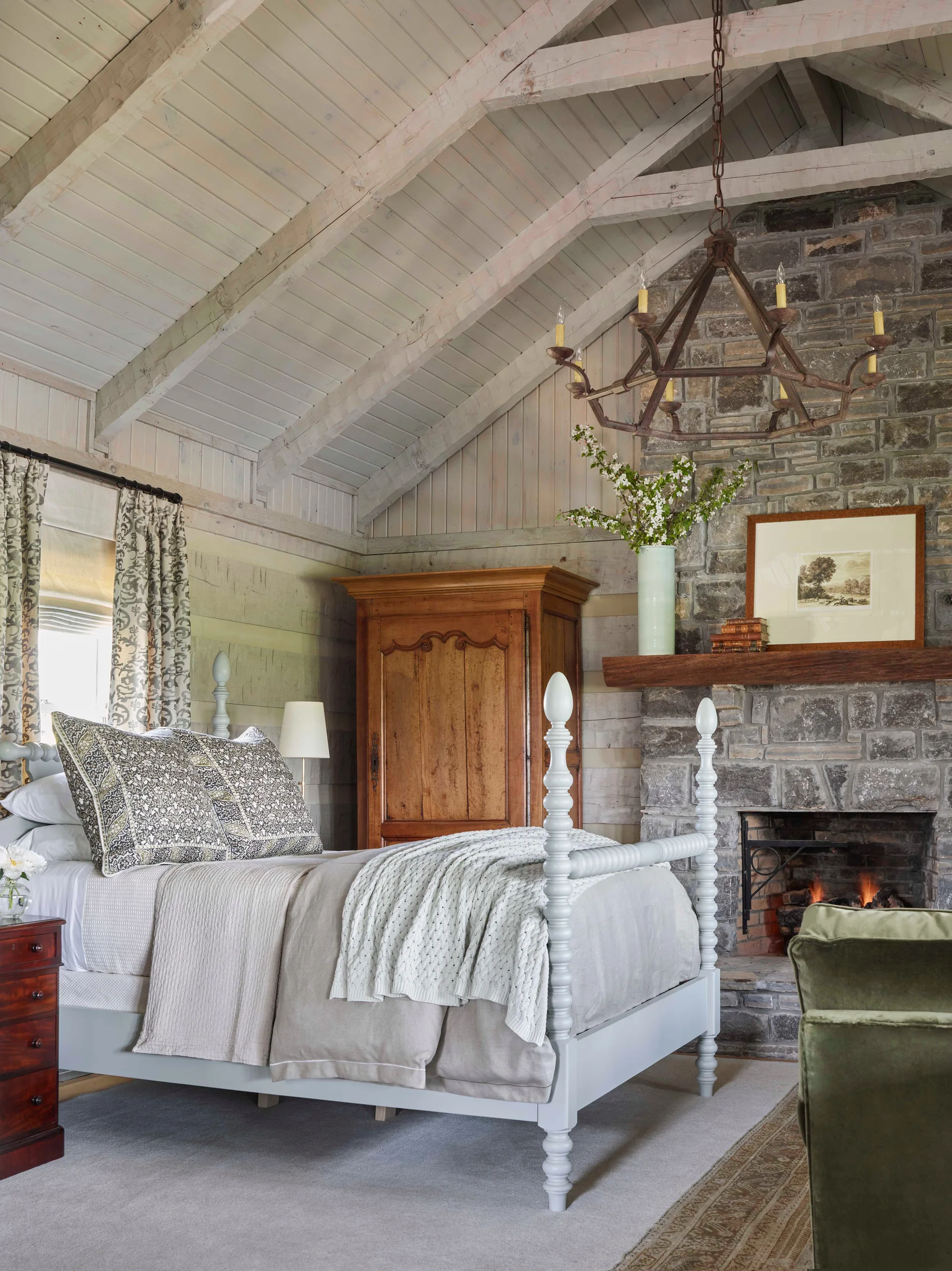 A rustic bedroom featuring a light blue spindle bed, a stone fireplace with a wood mantel, and a vaulted wood ceiling with exposed beams.