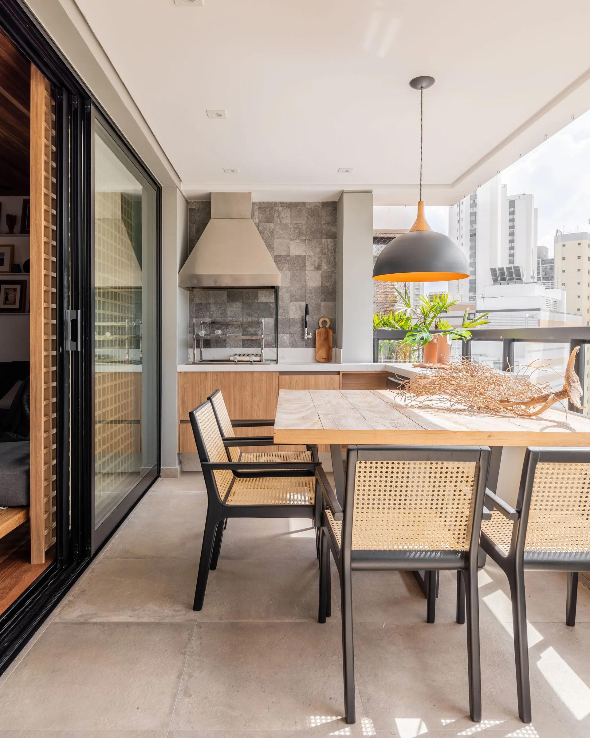 An urban balcony features a wooden dining table paired with black cane chairs next to an integrated outdoor kitchen with a stone backsplash.
