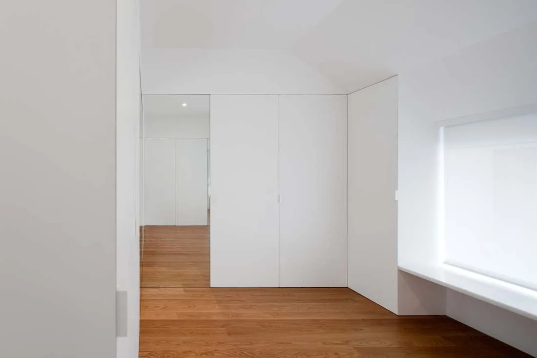 A minimalist room featuring white built-in cabinetry with flush doors alongside warm wood flooring and a recessed window.