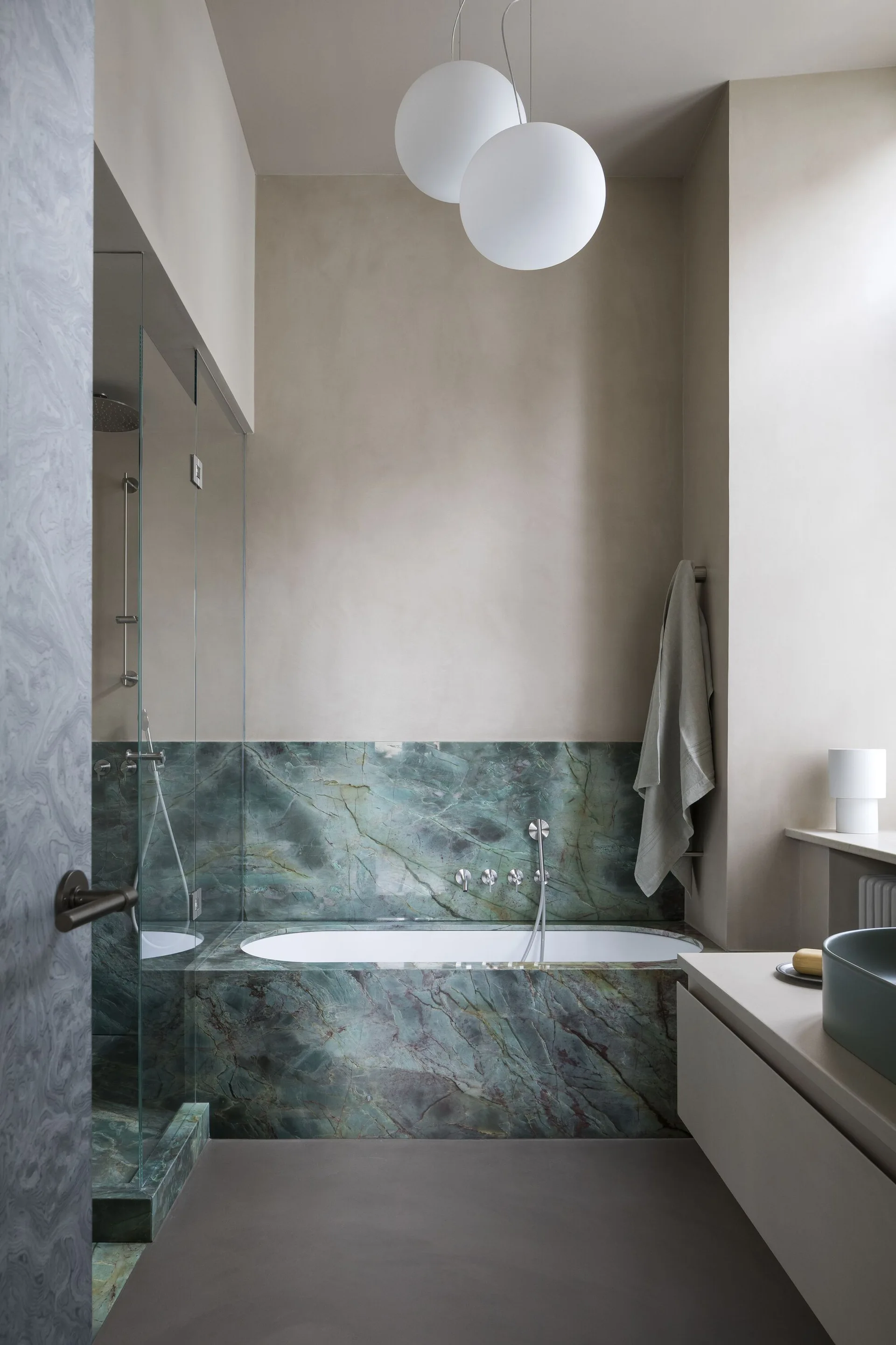 A modern bathroom features a built-in tub wrapped in green veined marble, neutral plaster walls, and two spherical pendant lights.
