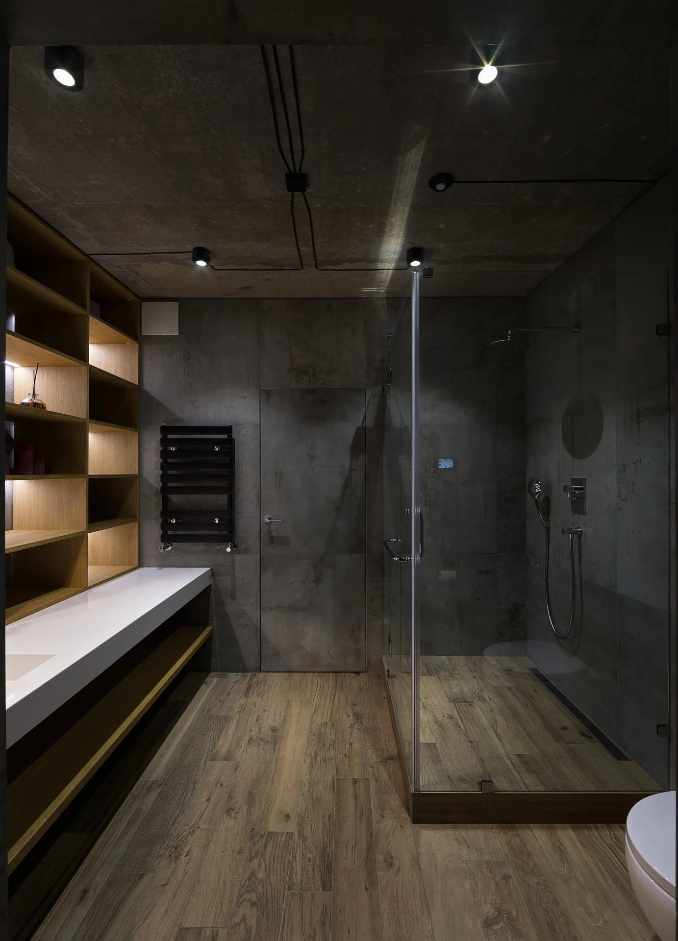 A modern industrial bathroom features grey concrete walls, a raw concrete ceiling, open wooden shelving with built-in lights, and a glass shower.