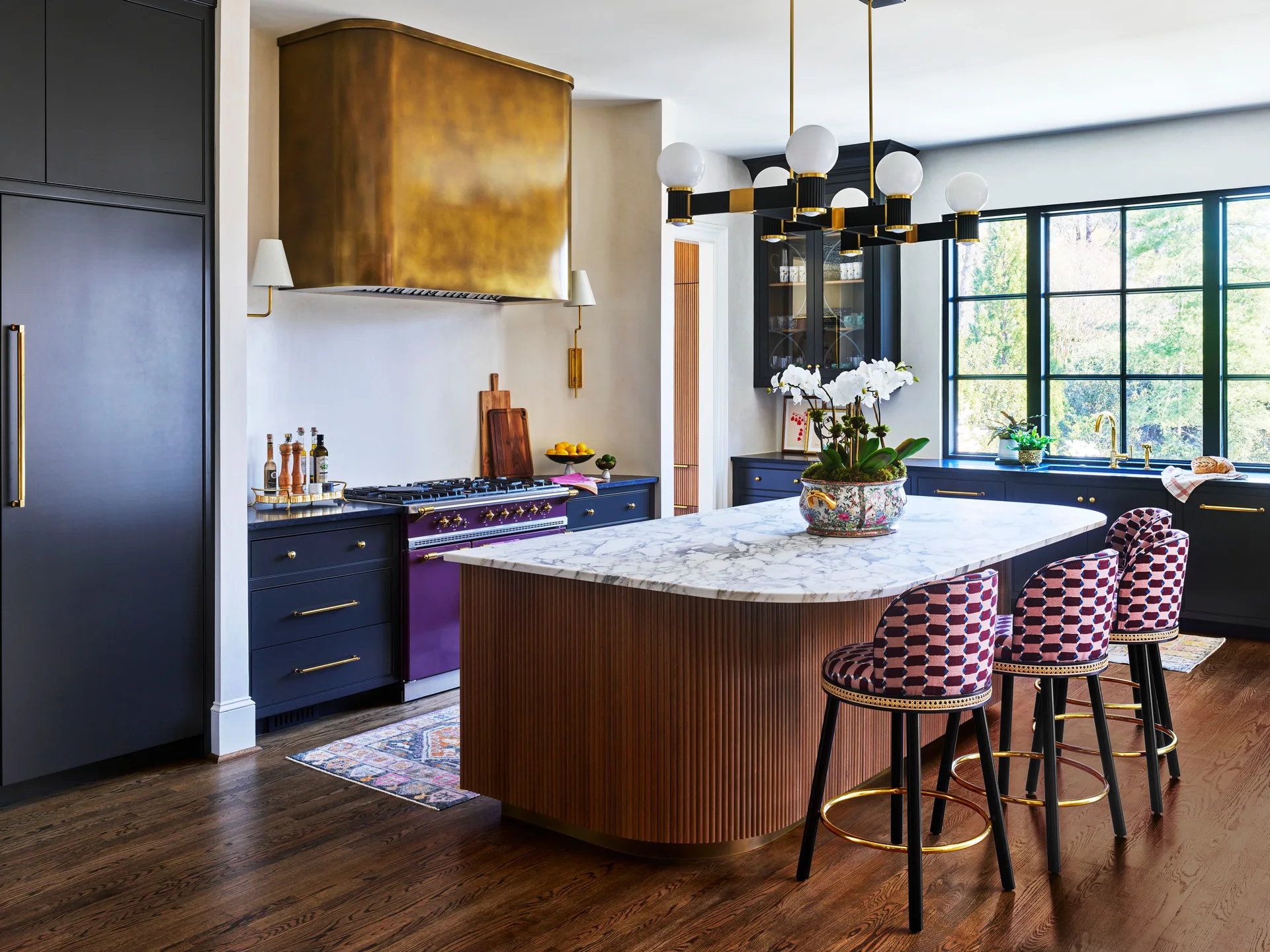 An eclectic kitchen features a fluted wood island with a marble top, dark navy cabinets, a purple stove, and a large brass range hood.