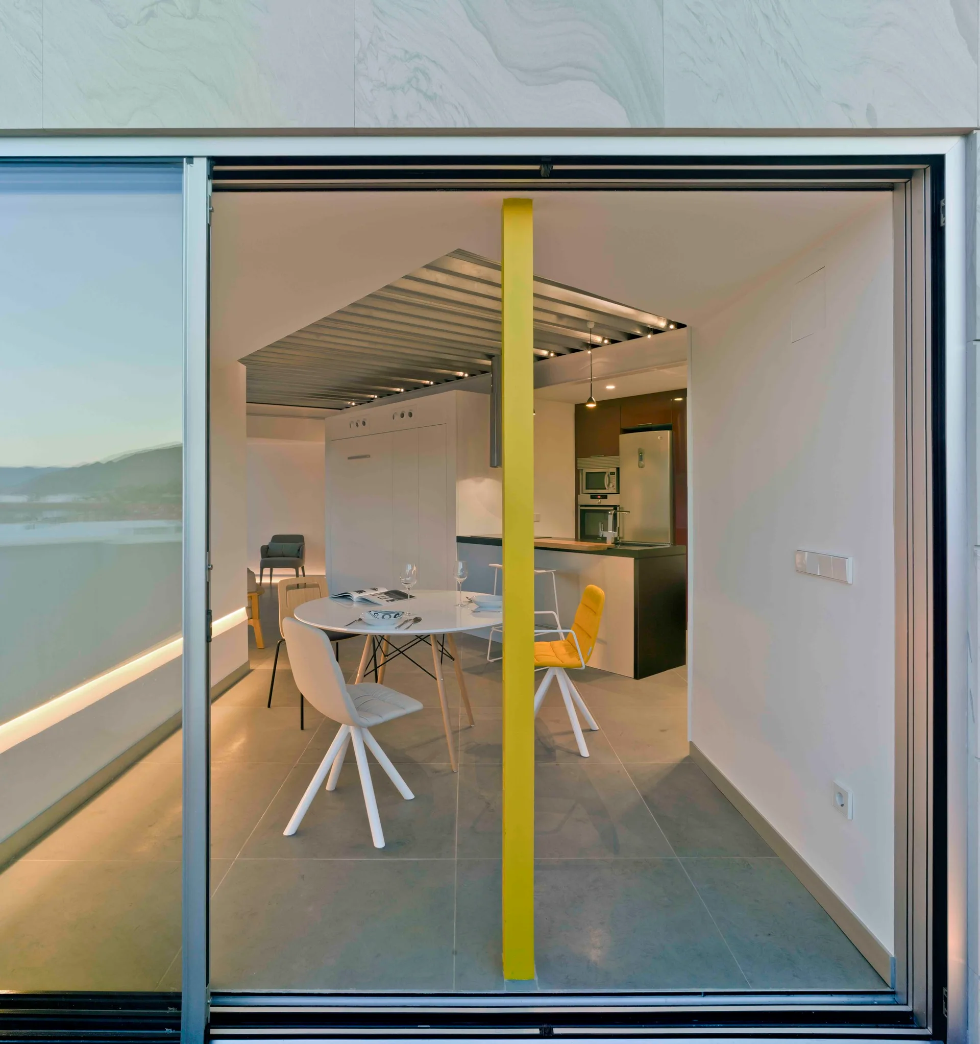 A modern open-concept dining room with a round white table, yellow chairs, and a bright yellow structural column visible through glass doors.