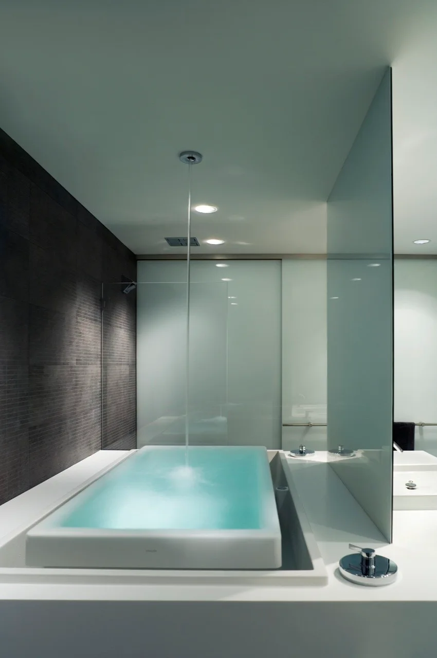 A modern minimalist bathroom featuring a built-in white bathtub filled with glowing water, a ceiling-fed faucet, and dark tiled walls.