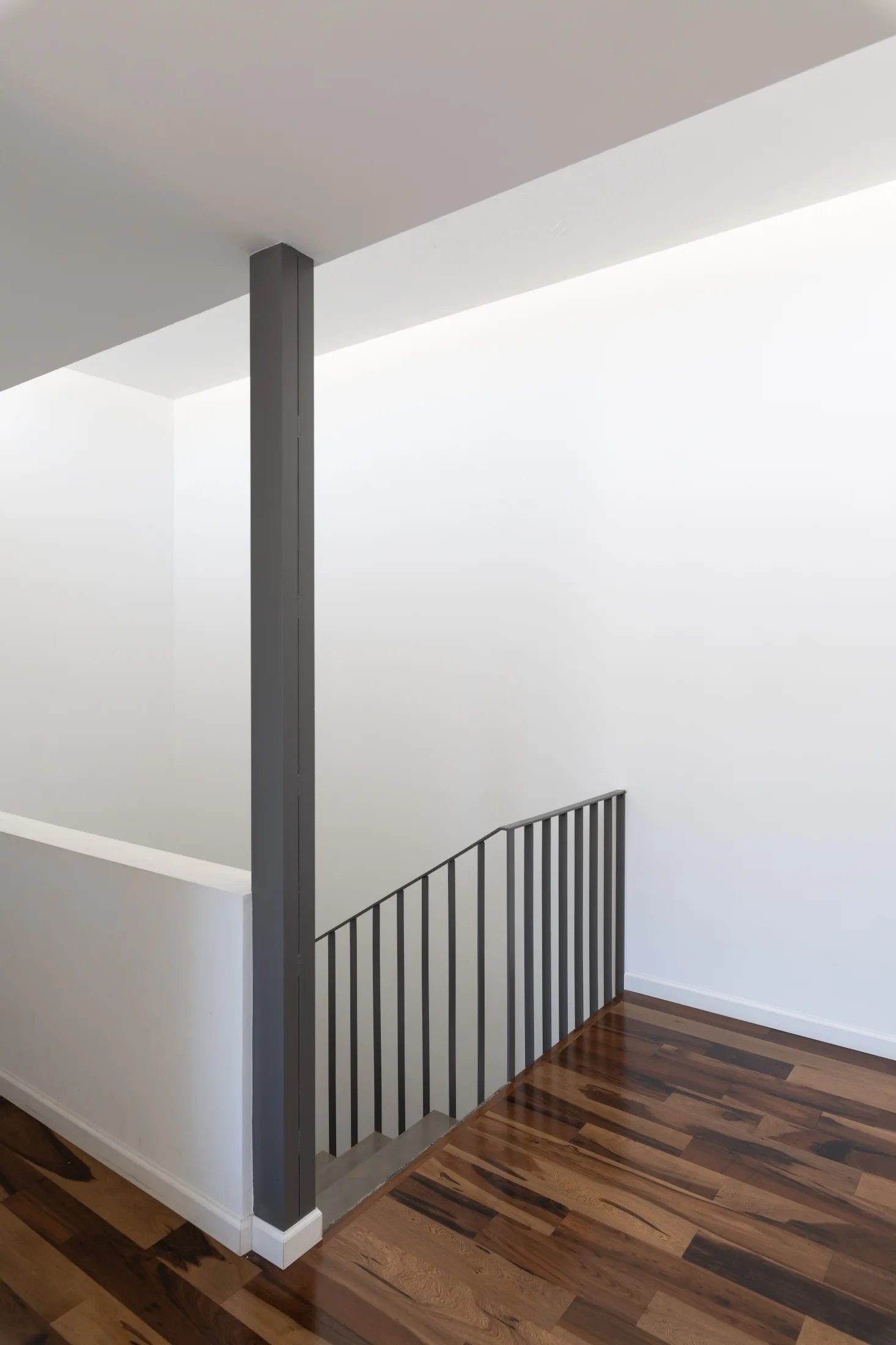 A minimalist stairwell features dark wood flooring, plain white walls, a dark grey support column, and a thin black metal railing.