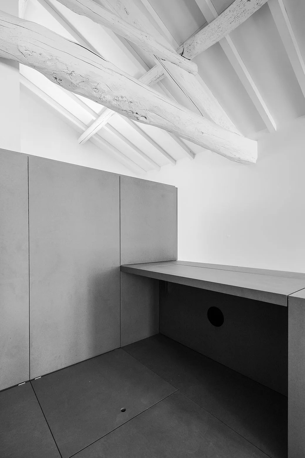 A minimalist gray stone-like built-in desk and matching wall panels sit beneath white painted exposed wood ceiling beams and a bright skylight.
