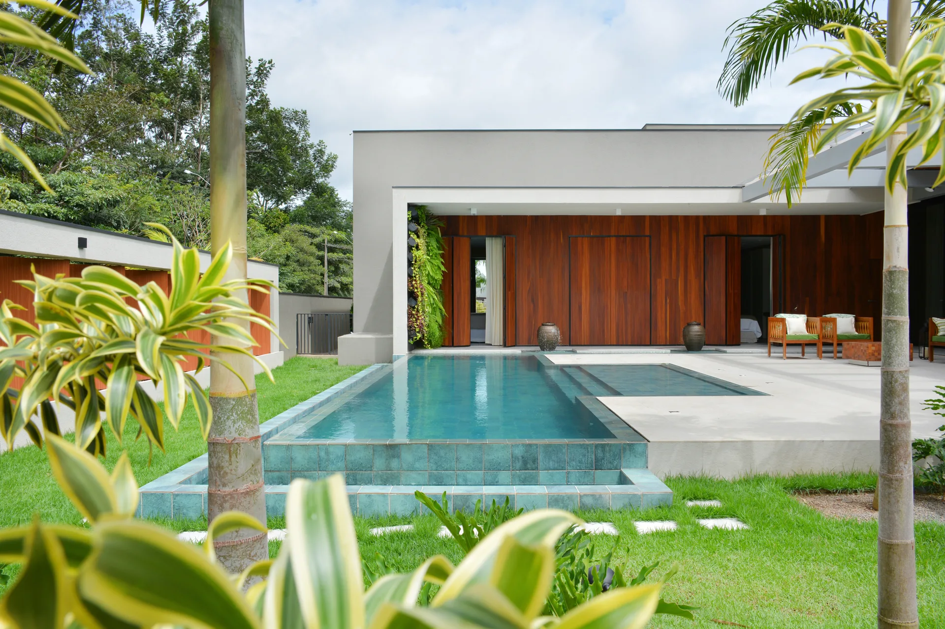 A modern rectangular swimming pool with teal tiling next to a concrete patio and a house with warm wood paneling.