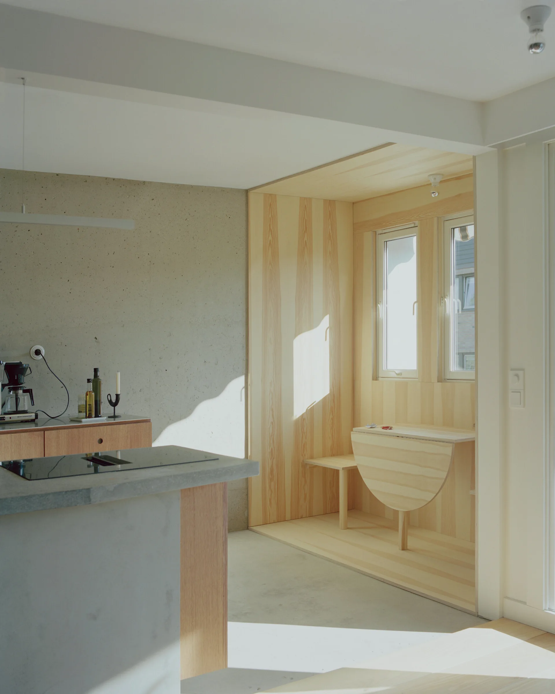 A minimalist kitchen features raw concrete walls, a concrete counter, and a built-in wooden dining nook with a drop-leaf table.