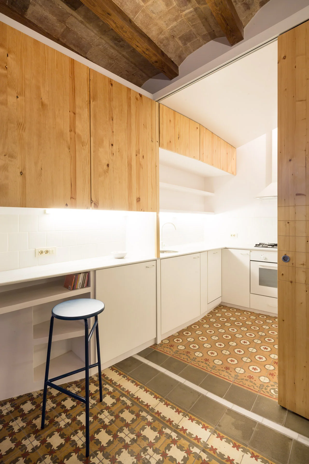 A contemporary kitchen featuring natural wood upper cabinets, white lower cabinets, patterned floor tiles, and an exposed brick vaulted ceiling.
