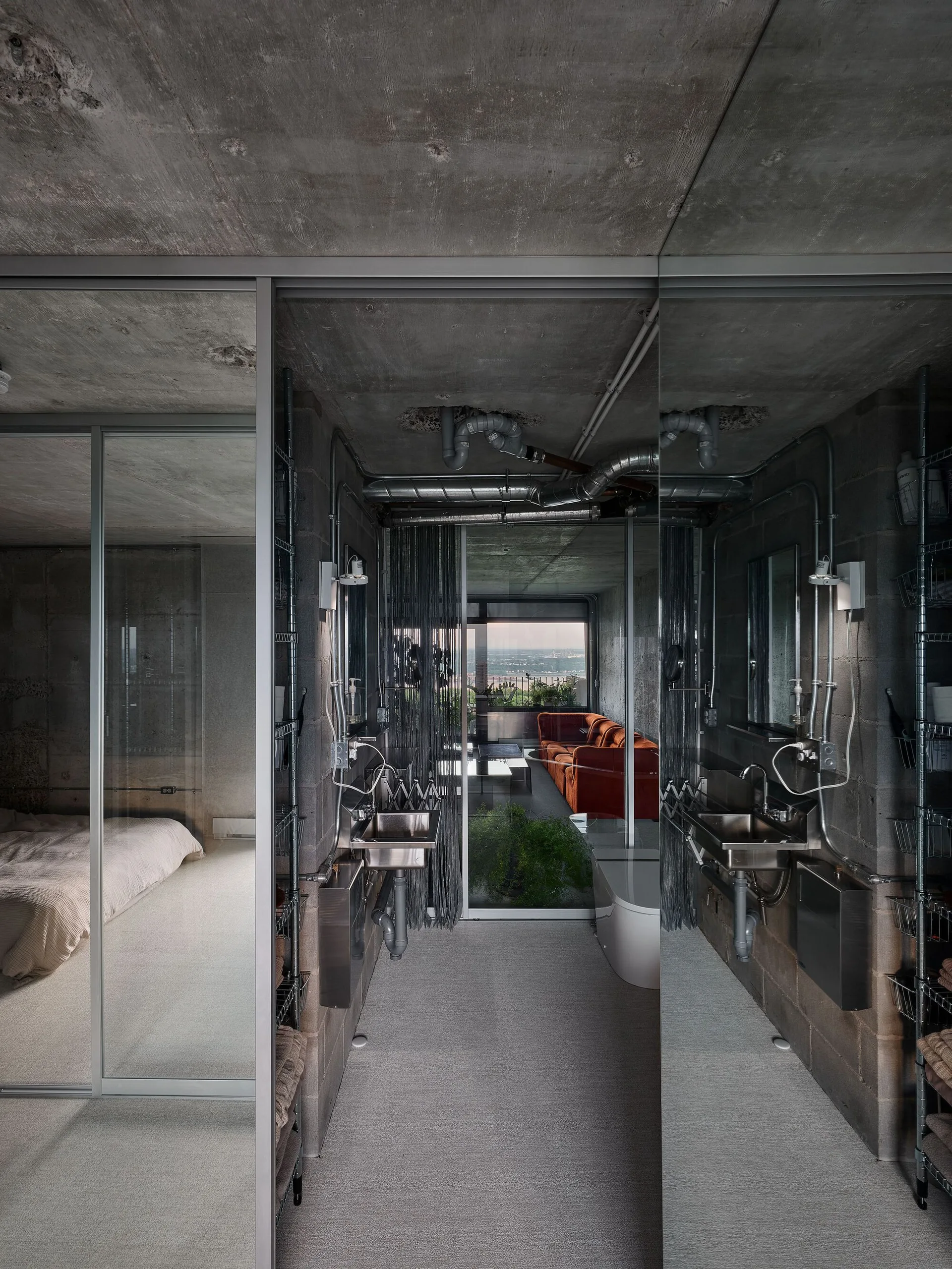 An industrial bathroom features raw concrete walls, exposed piping, a stainless steel sink, and floor-to-ceiling mirrored panels.