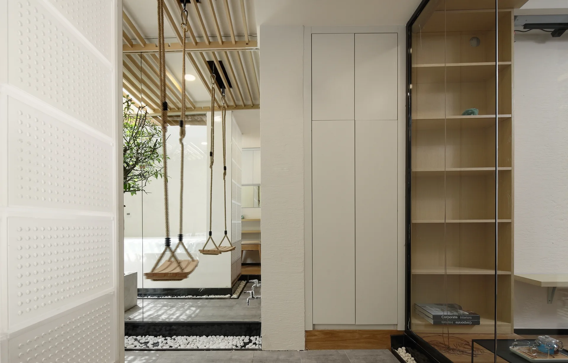 Two wooden swings hang from a slatted ceiling behind a glass partition in a minimalist room with white cabinets and wood shelving.