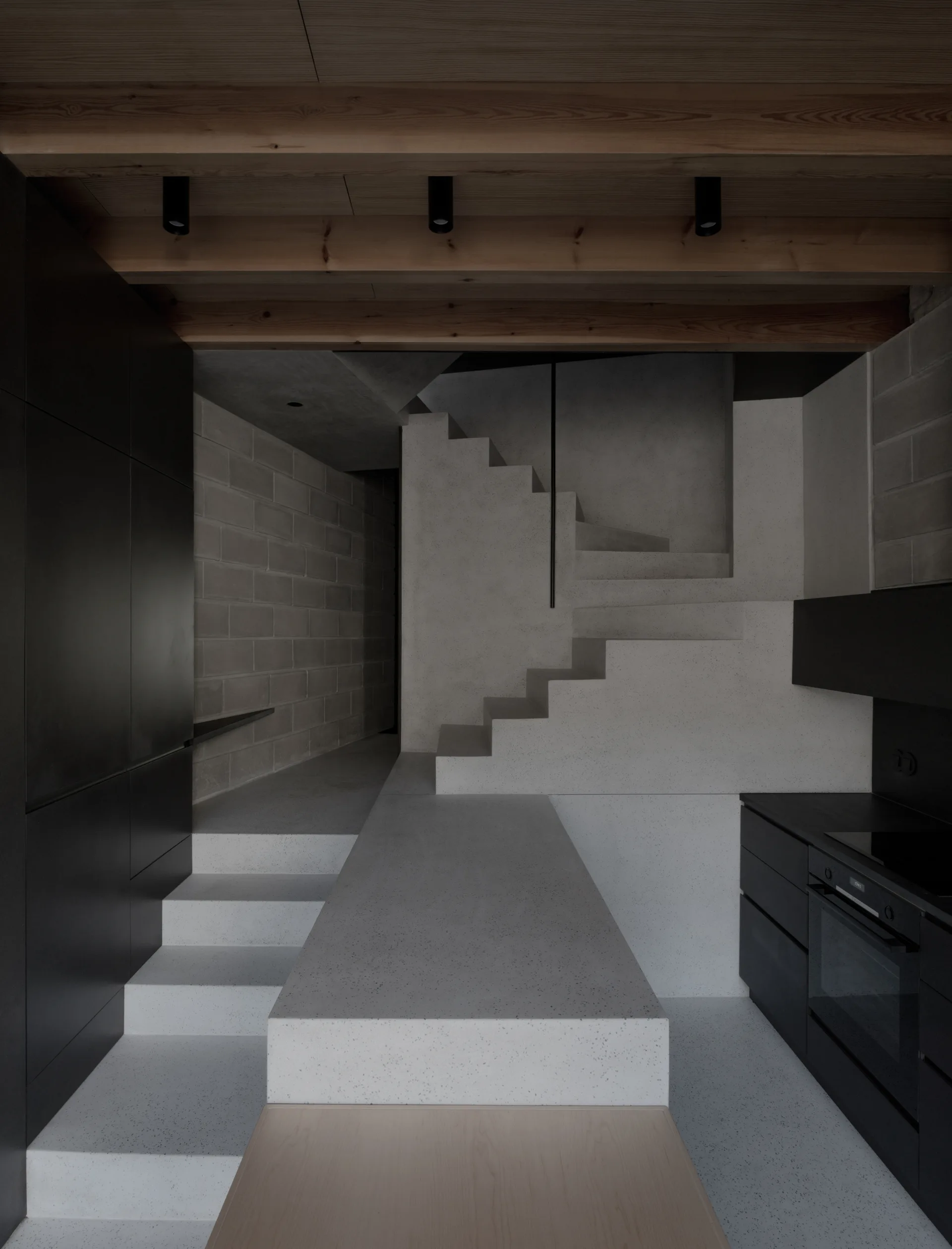 A minimalist interior featuring a concrete staircase, matte black kitchen cabinets, and exposed wooden ceiling beams with recessed spot lights.