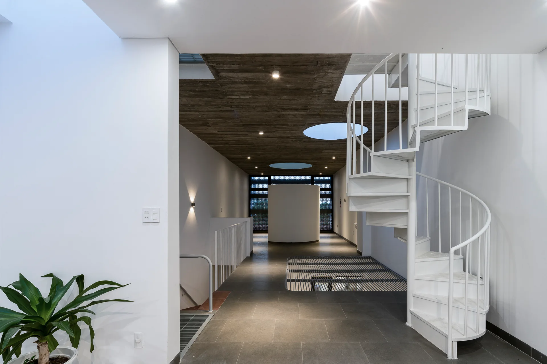 A modern hallway features a white spiral staircase next to circular skylights cut into a rough concrete ceiling and a dark grey tiled floor.