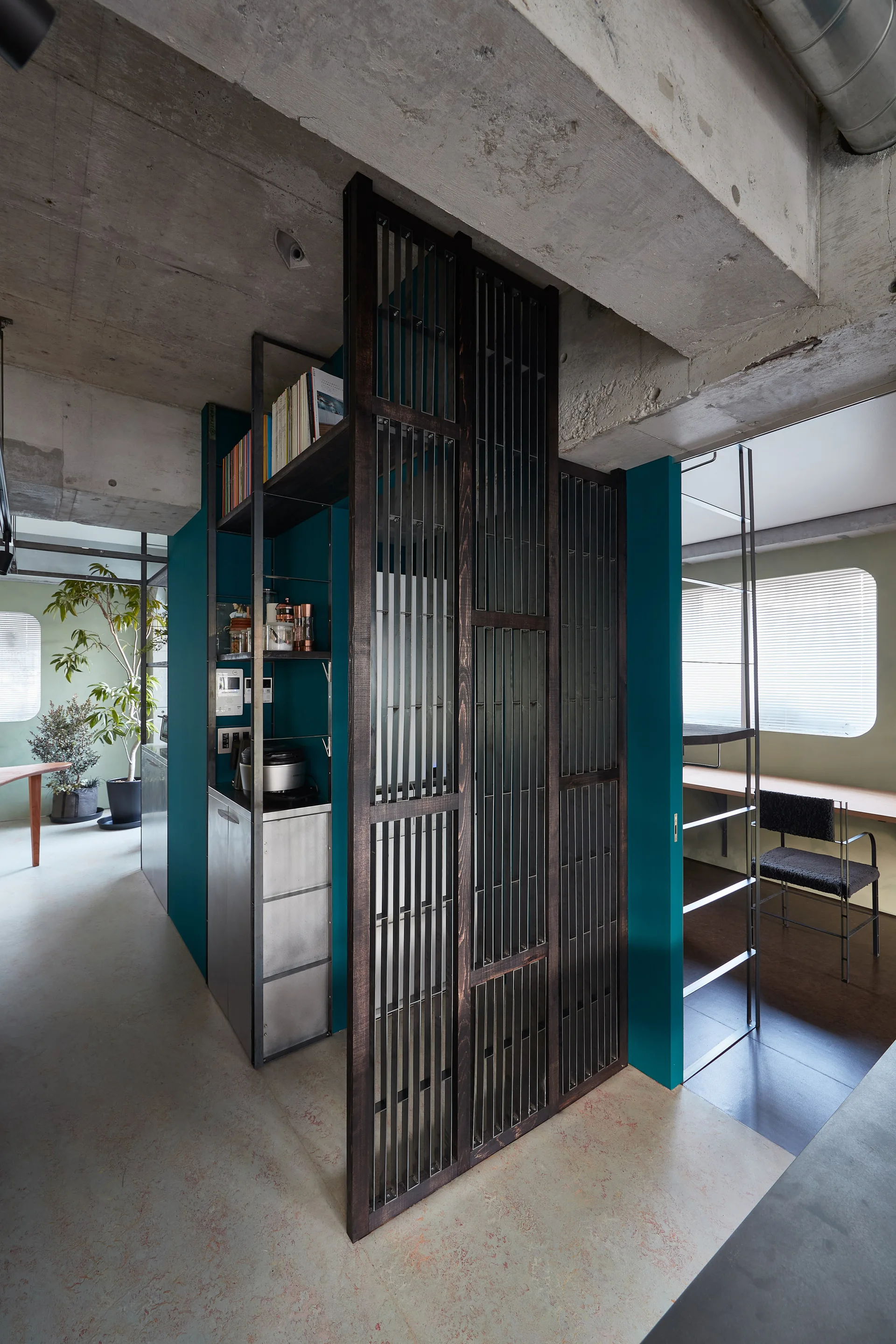 A dark slatted room partition stands between a kitchen with teal walls and a study nook beneath an exposed concrete ceiling.