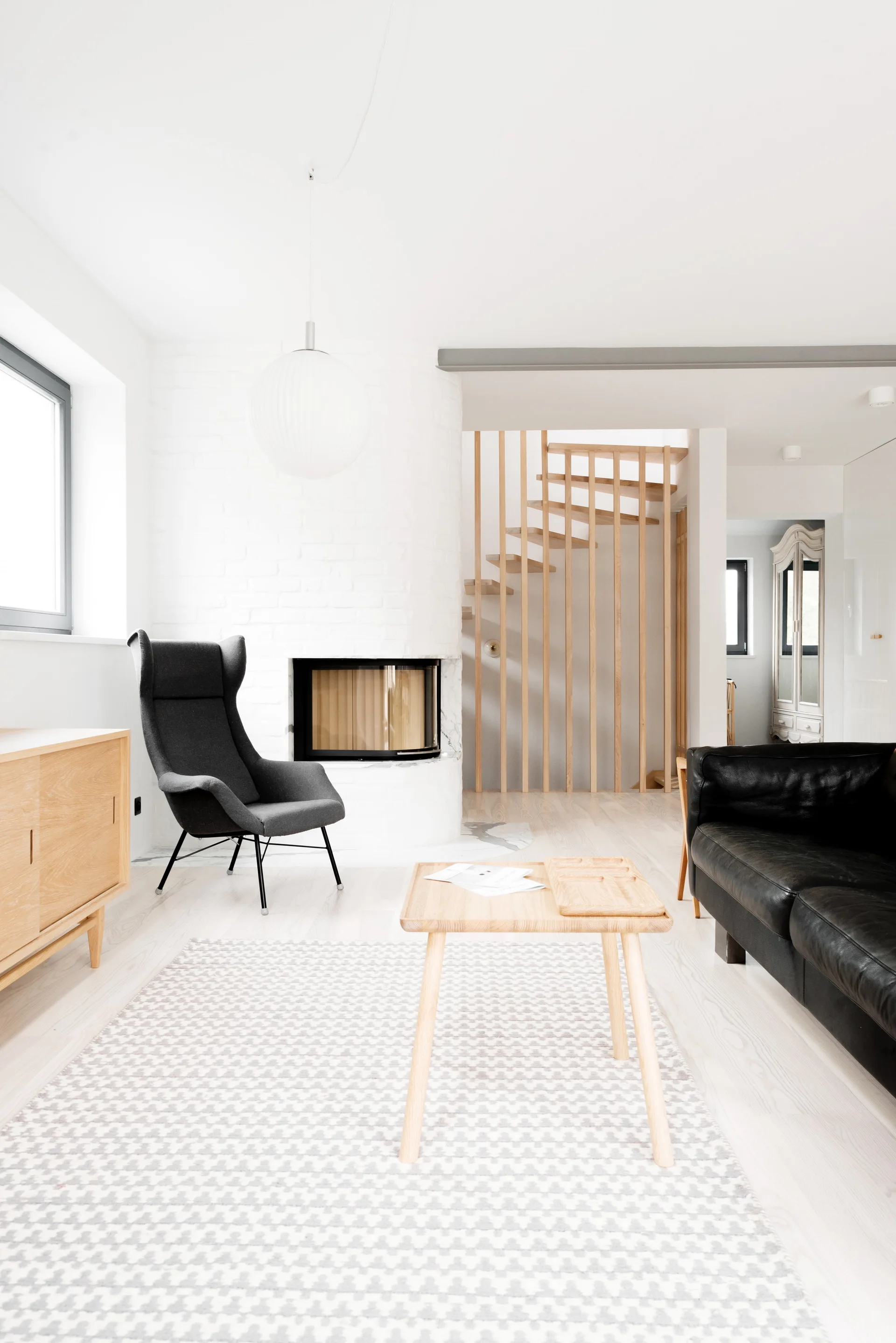 A bright living room features a curved white brick fireplace, a dark grey armchair, a light wood coffee table, and a slatted wooden staircase.