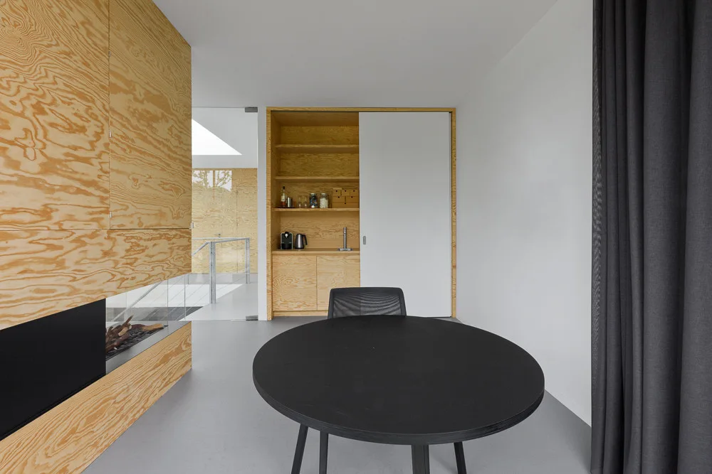 A circular black table sits in a minimalist room with warm plywood wall paneling, built-in storage with a white sliding door, and dark curtains.