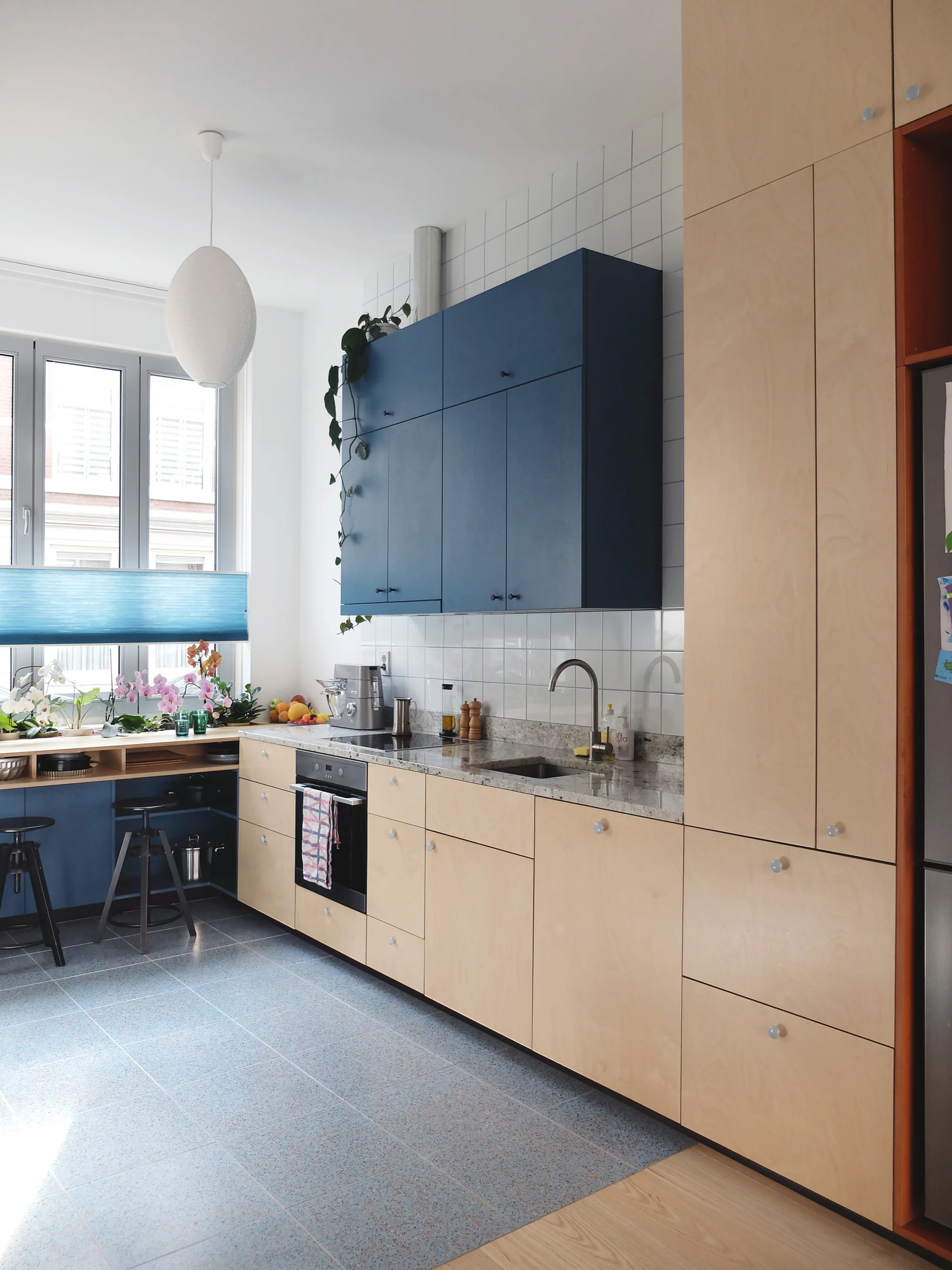 A modern kitchen features light wood plywood base cabinets, deep blue upper cabinets, a white tiled backsplash, and a blue speckled floor.