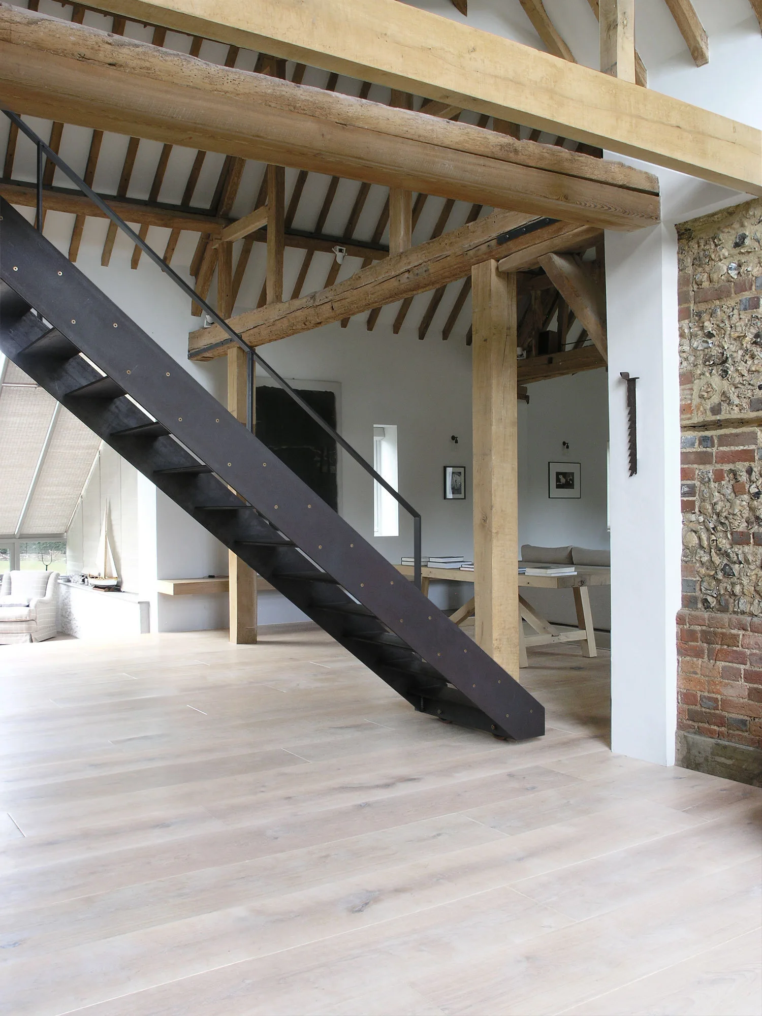 A black metal open-tread staircase stands in a spacious room with light wood floors, exposed timber ceiling beams, and a brick-and-stone wall segment.