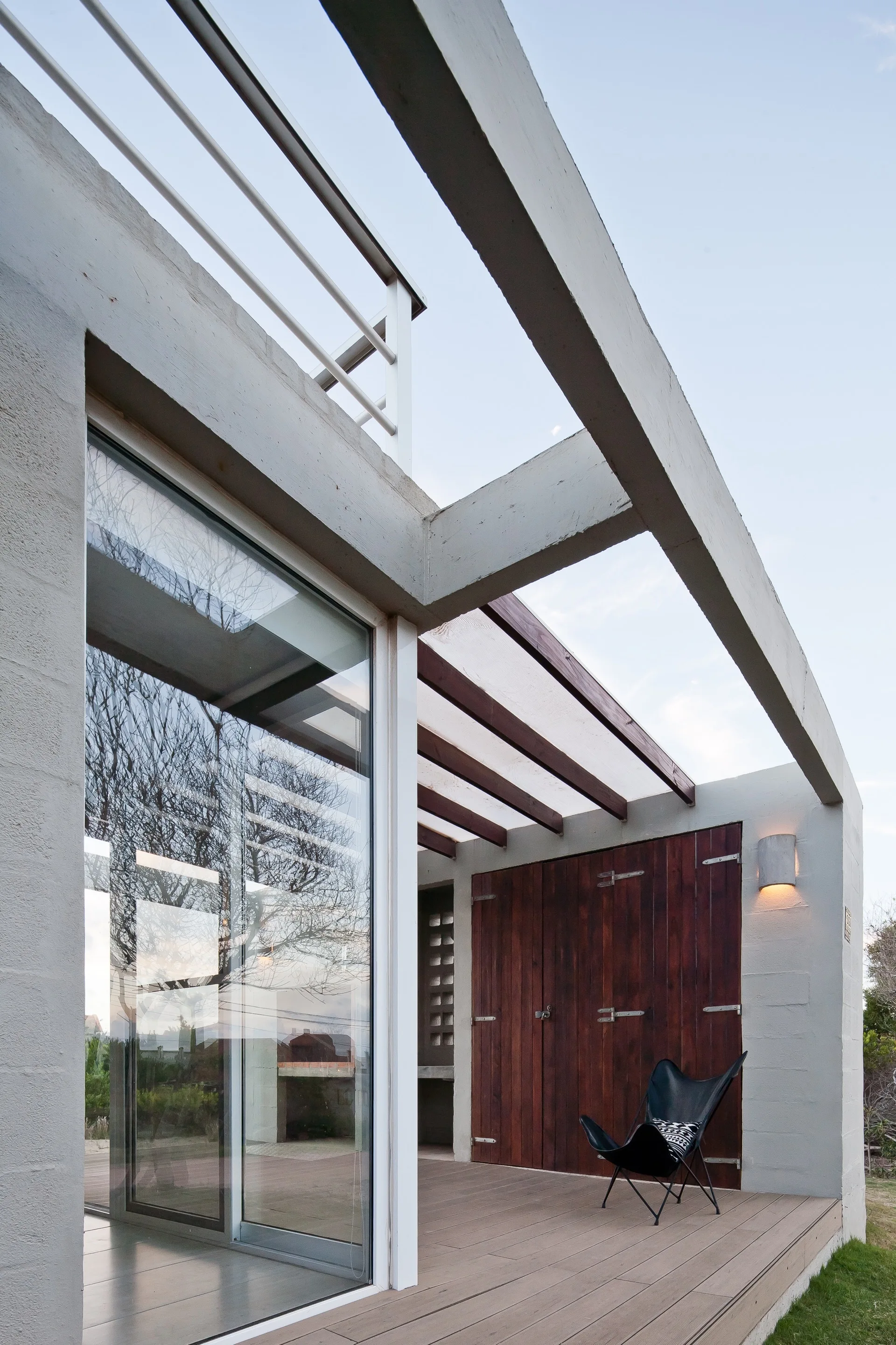 A black butterfly chair sits on a wooden deck patio beneath structural concrete beams next to a dark wood panel door and glass sliding door.