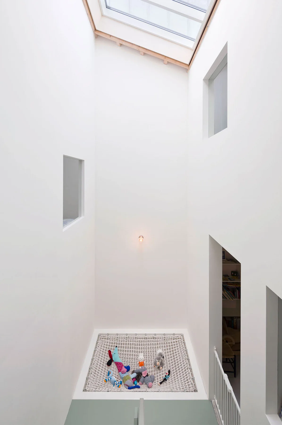 A tall white minimalist atrium features a suspended rope net floor holding stuffed animals beneath a bright overhead skylight.