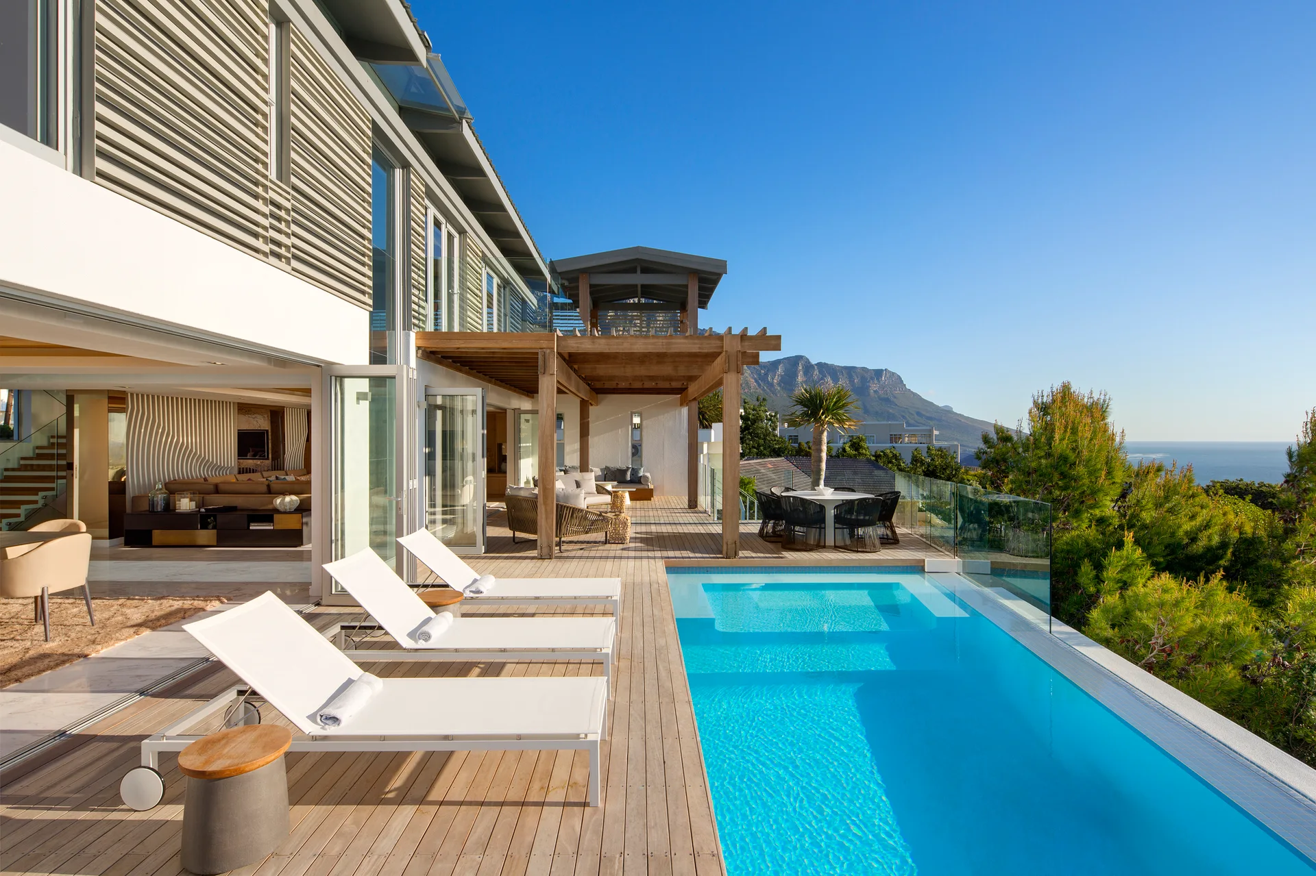 White sun loungers sit on a wide wooden deck next to a bright blue swimming pool, with a wooden pergola and mountain views in the background.