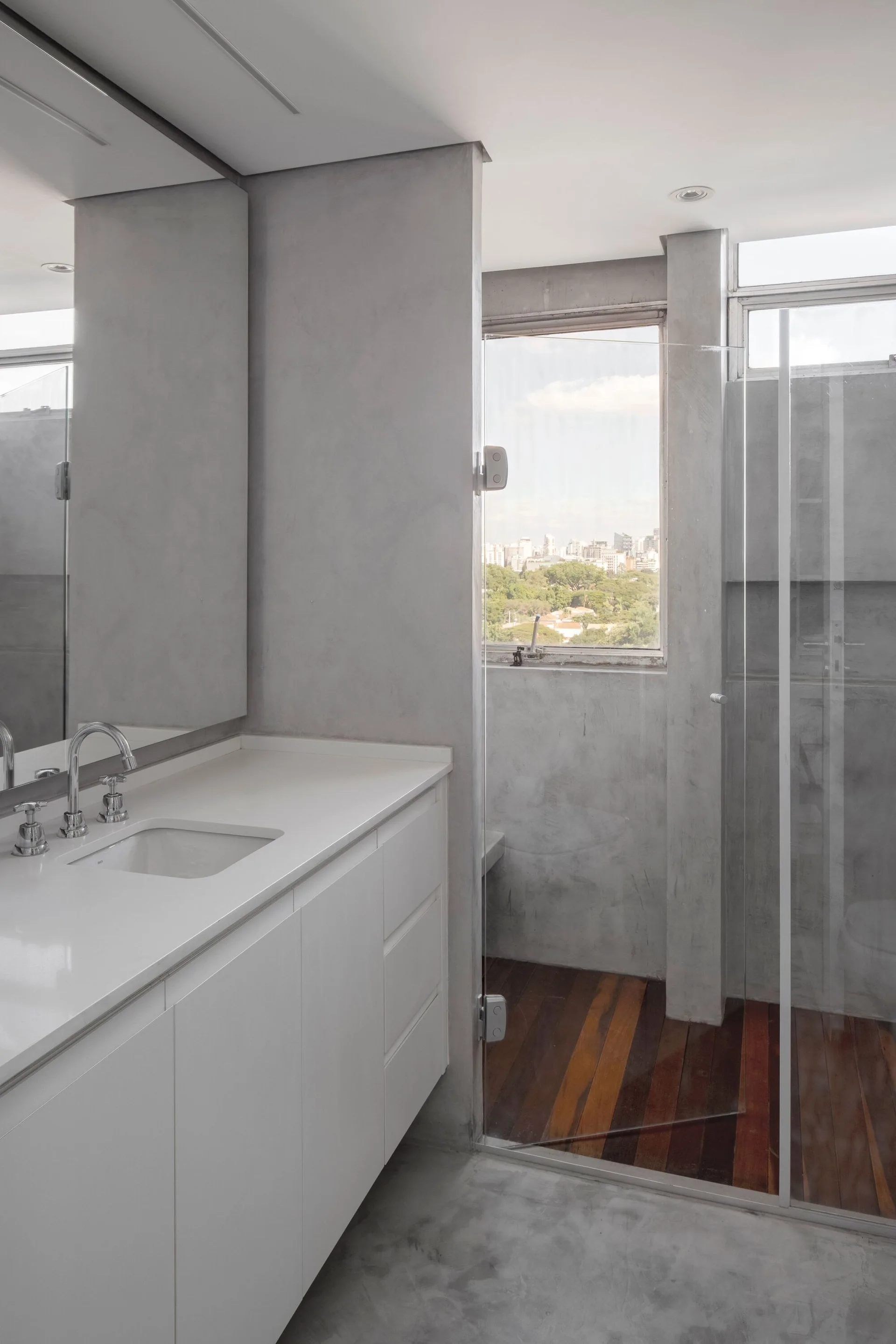A minimalist gray concrete bathroom featuring a white vanity with integrated sink and a glass-enclosed shower with wood plank flooring.