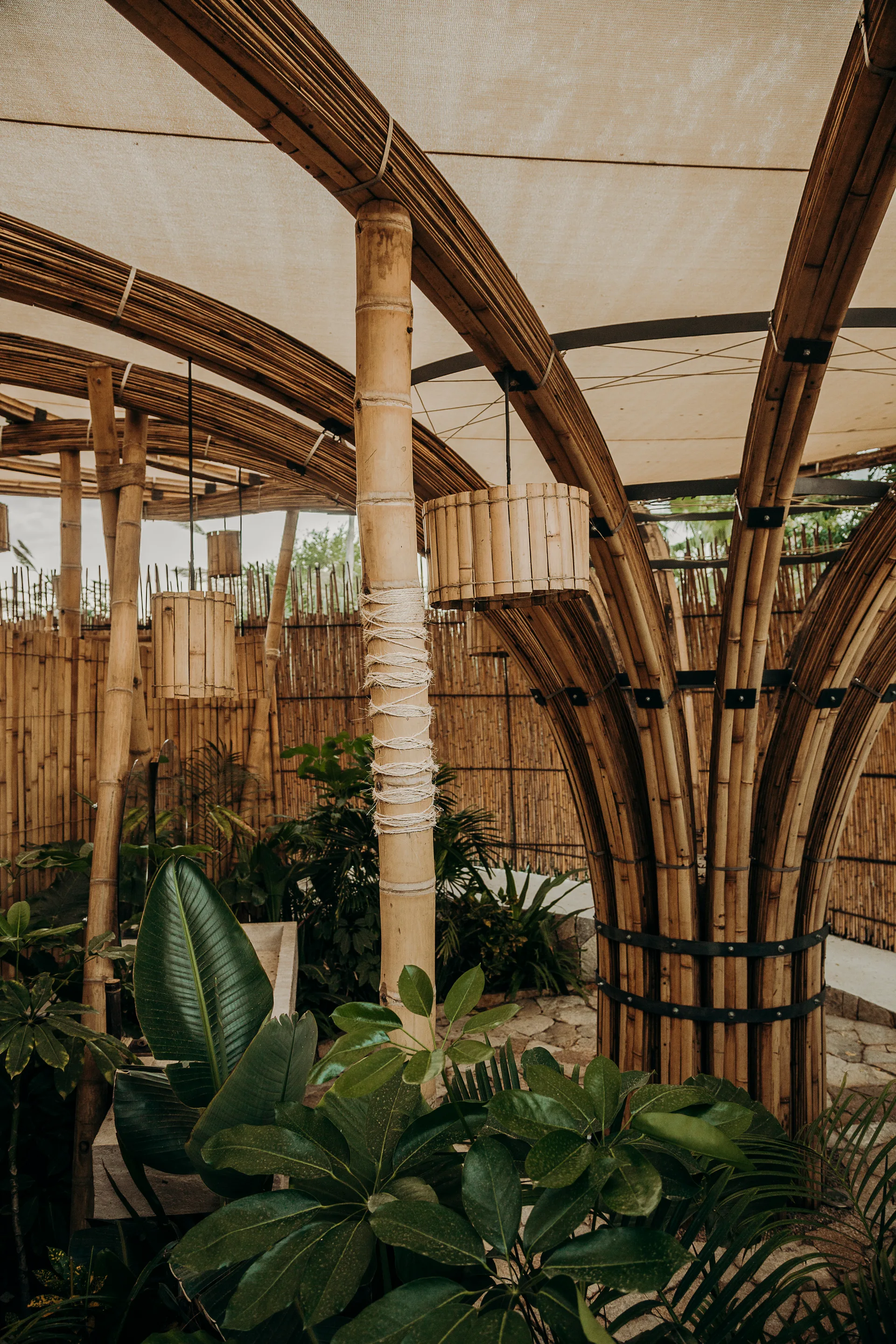 An outdoor patio features curved bamboo columns supporting a fabric canopy shade above tropical green plants and a slatted bamboo screen.