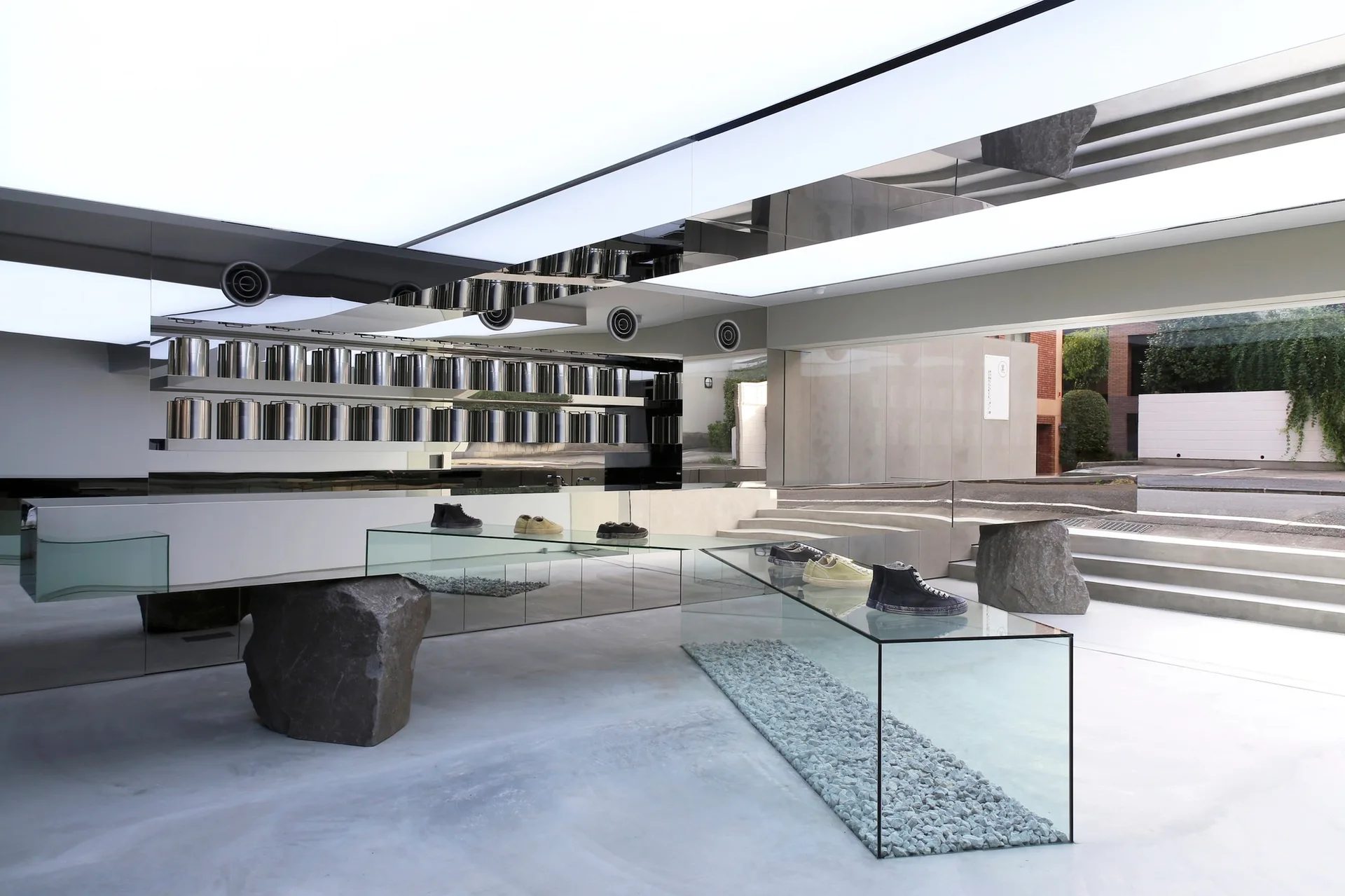 A minimalist retail store interior features glass display tables supported by large stone boulders, a mirrored wall, and an illuminated ceiling.