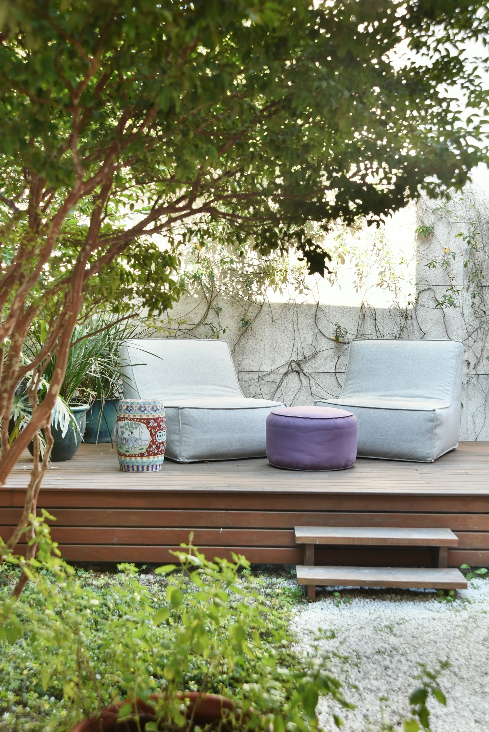 Two gray lounge chairs sit on a low wooden deck in a leafy garden patio with a purple ottoman and a ceramic garden stool.