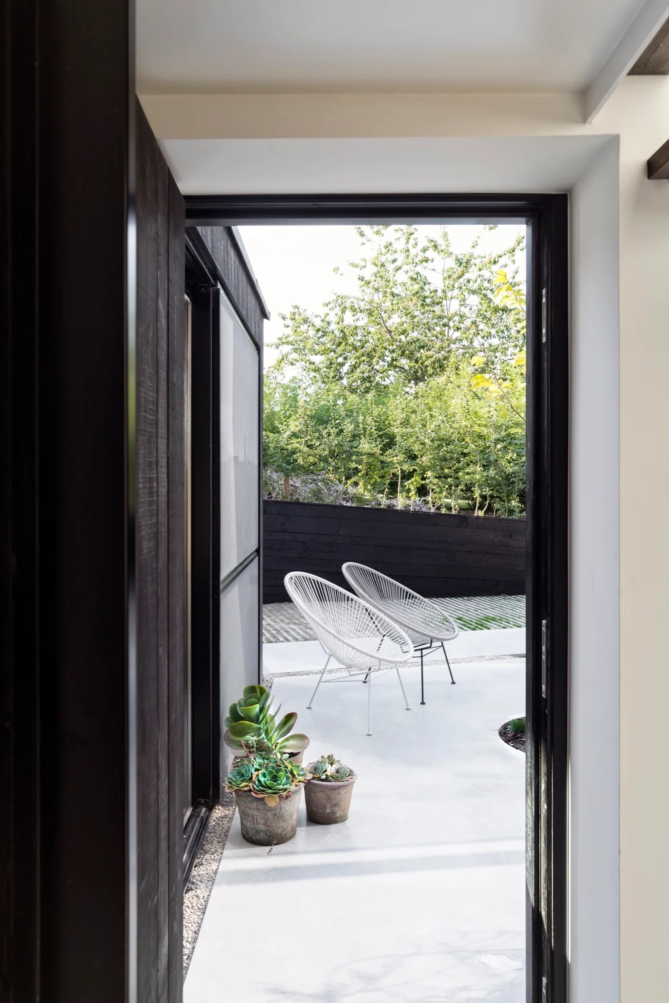 A view through a dark doorway onto a concrete patio with two white woven lounge chairs and potted succulents.