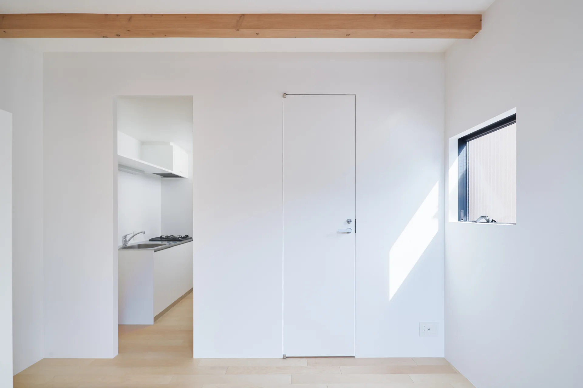 A minimalist white hallway features a flush door, a rectangular window, and a raw wood ceiling beam, with an open doorway leading to a small kitchen.