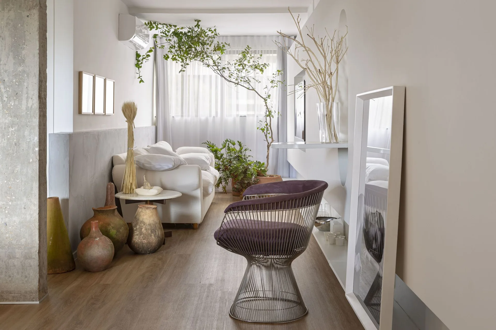 A narrow living room features a white sofa, a wire armchair with a purple seat, and large clay pots on a wood floor.