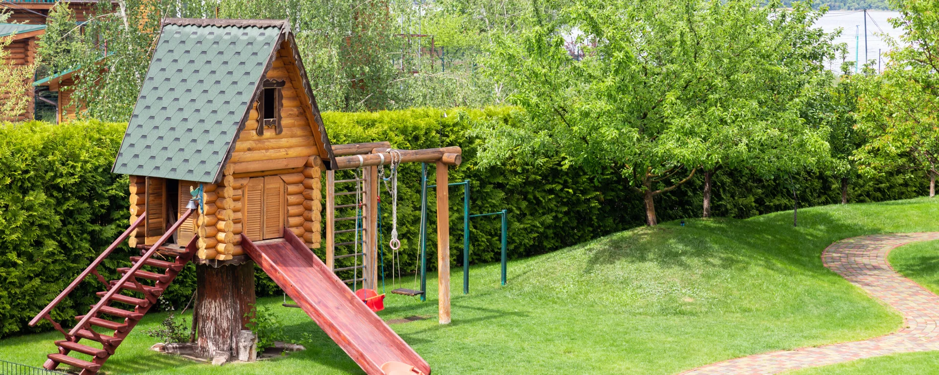 A children's log cabin playhouse with a red slide and a swing set stands on a bright green lawn.
