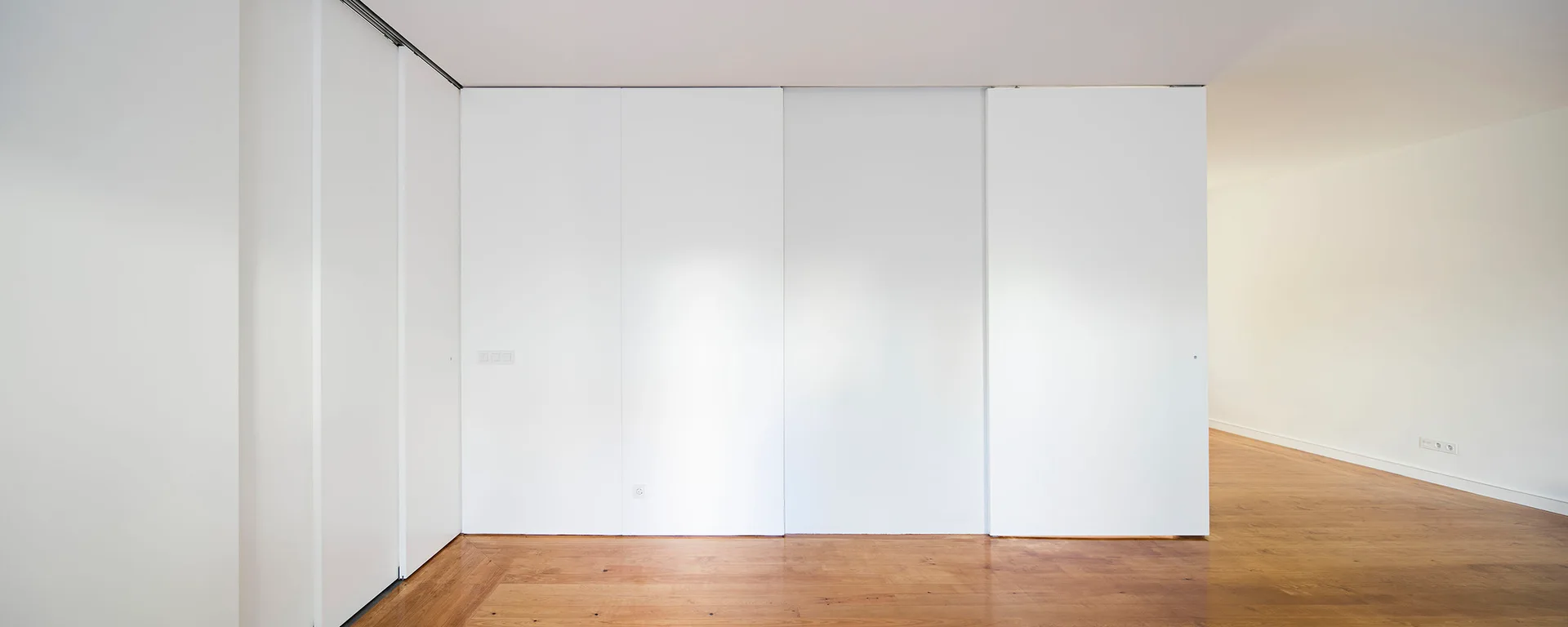 Seamless floor-to-ceiling white cabinet panels line a minimalist room with warm wood plank flooring.