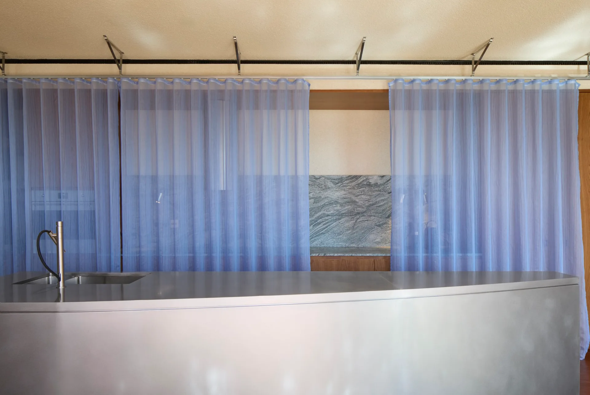 A stainless steel kitchen island with a faucet stands before a stone-veined backsplash partially obscured by hanging sheer blue partition curtains.