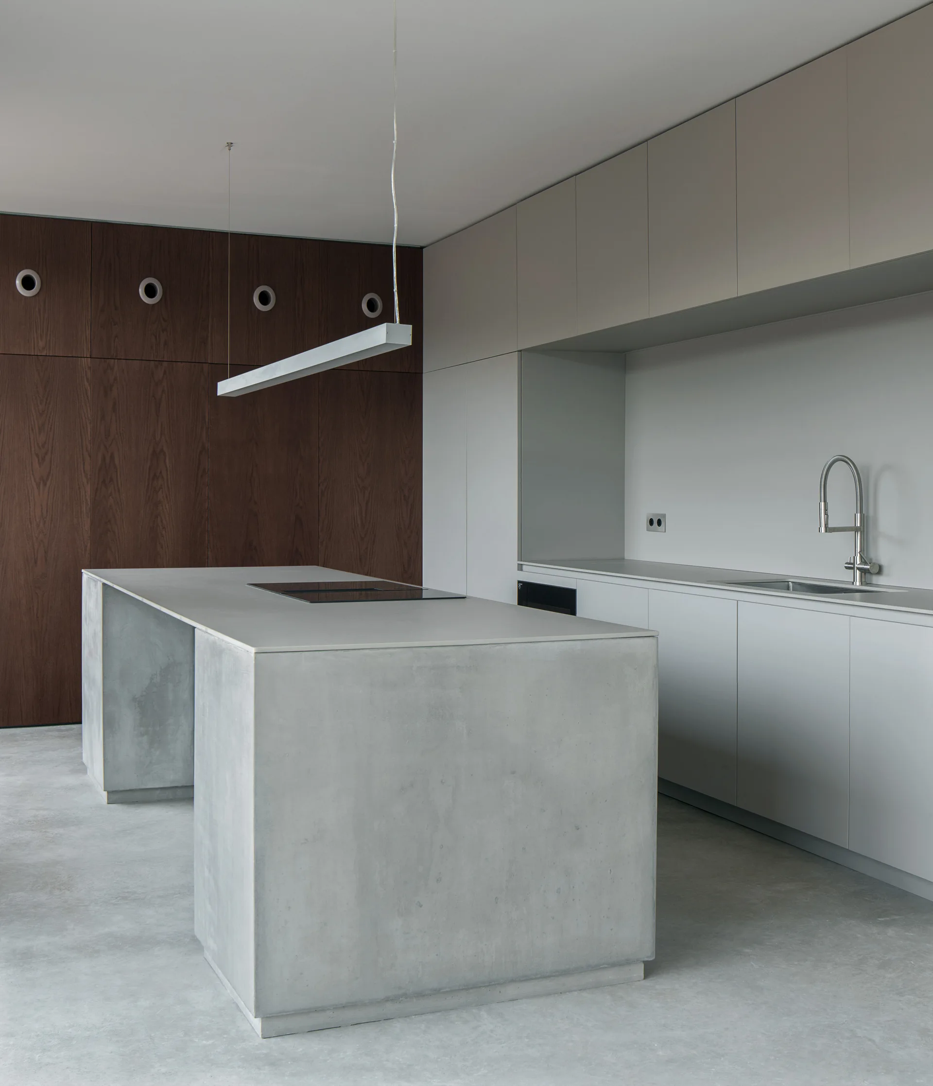A minimalist kitchen features a large concrete island, light grey handleless cabinets, a dark wood-paneled wall, and a linear pendant light.
