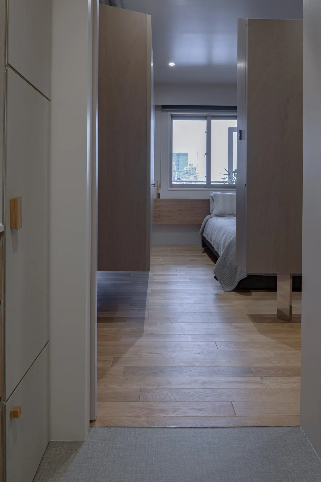 A view through a doorway into a minimalist bedroom featuring light wood flooring, tall wooden partition wardrobes, and a low bed near a window.