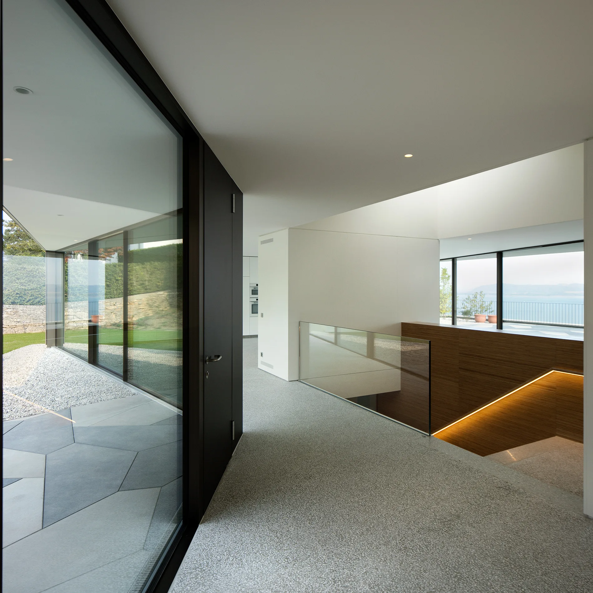 A modern hallway features speckled terrazzo flooring, floor-to-ceiling glass walls, and a wood-paneled staircase with integrated strip lighting.