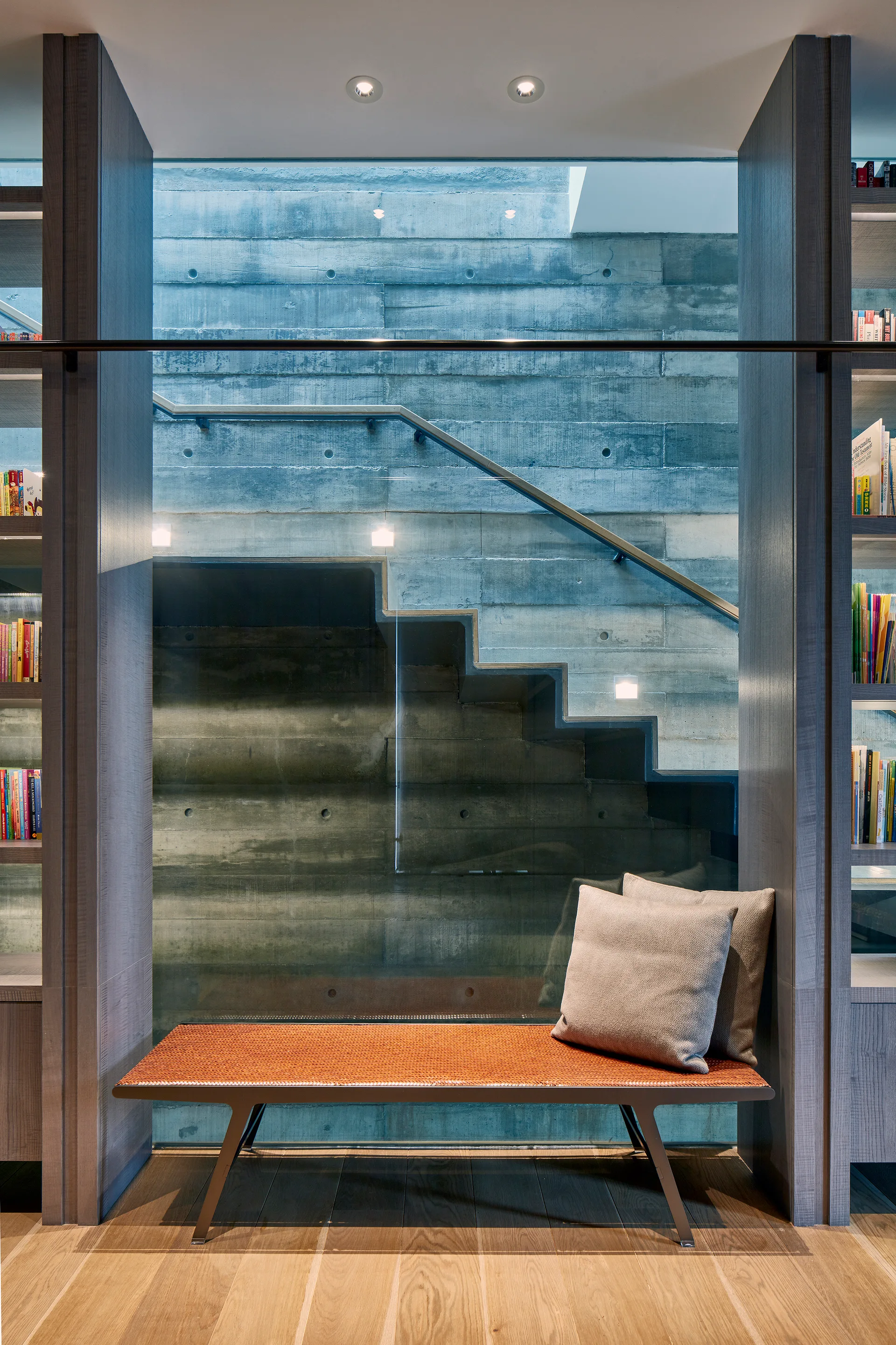 An orange woven bench sits on a wood floor between two wooden bookshelves, positioned in front of a glass partition showing a concrete staircase.