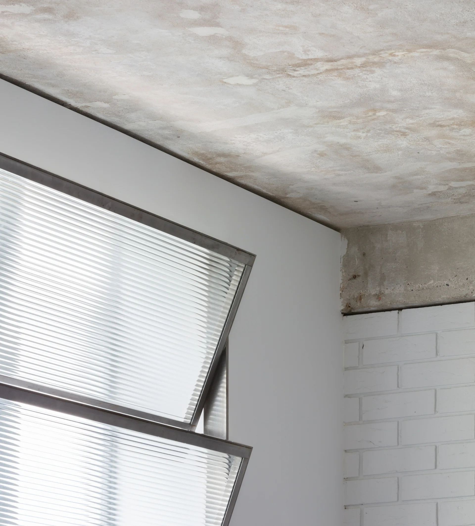 An architectural detail showing an open hinged window with ribbed glass set against a white wall, a painted brick wall, and a raw concrete ceiling.