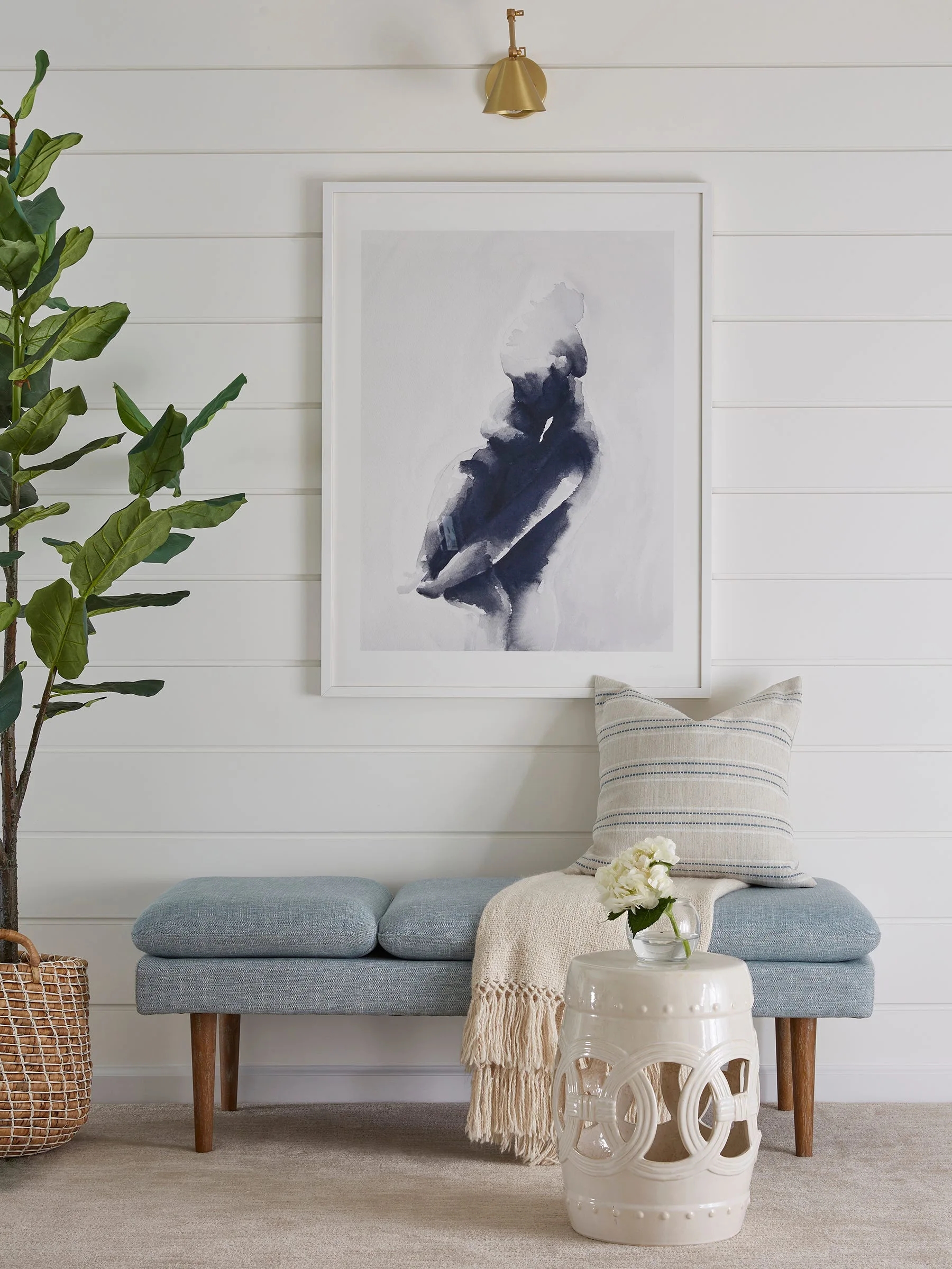A light blue upholstered bench with a striped cushion sits against a white shiplap wall beneath a framed abstract painting and a brass sconce.