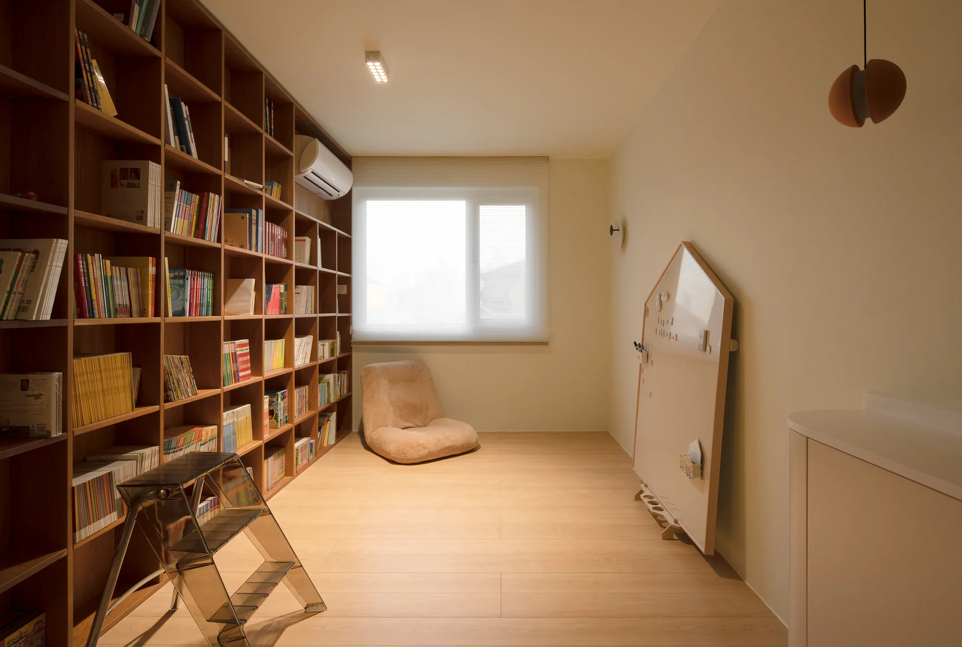 A minimalist study room features a full wall of wooden bookshelves filled with books, a low beige floor chair, and a house-shaped whiteboard.