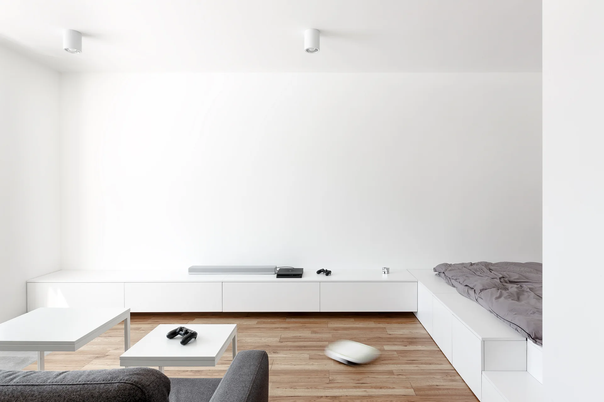 A minimalist room featuring a long white built-in cabinet that transitions into a platform bed, set against white walls and light wood flooring.