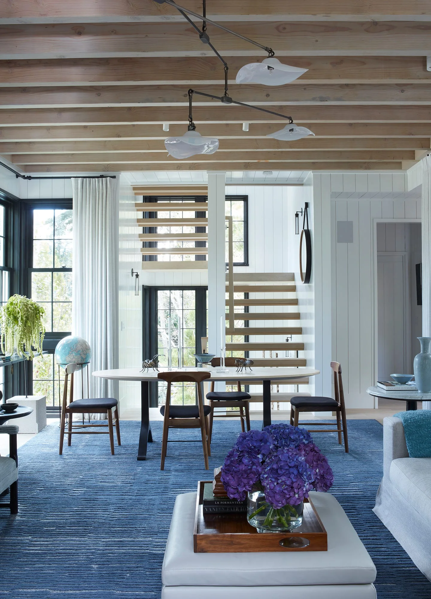 An open dining area features an oval wood table on a blue ribbed rug, exposed ceiling beams, and a floating wood staircase.