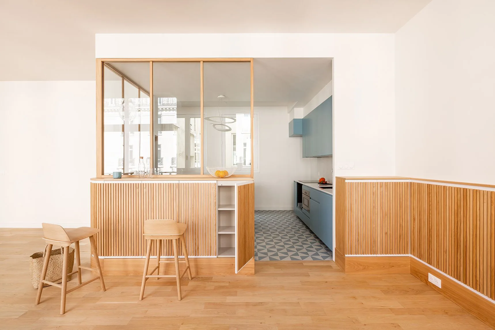 A slatted wood partition with glass panels separates a living space from a kitchen with muted blue cabinets and geometric floor tiles.