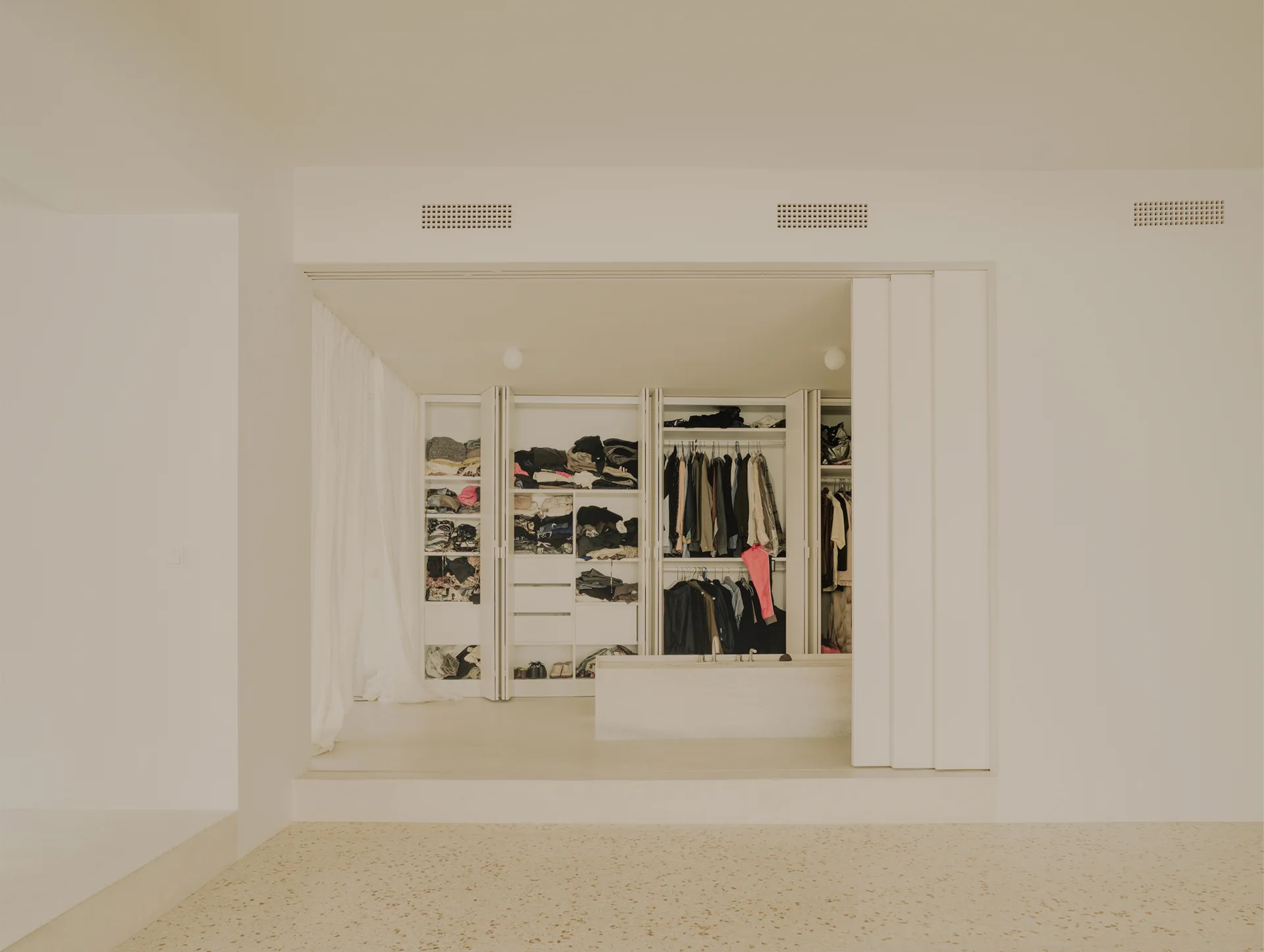 A minimalist walk-in closet system on a raised platform with white built-in wardrobes, a sheer curtain, and speckled terrazzo flooring.