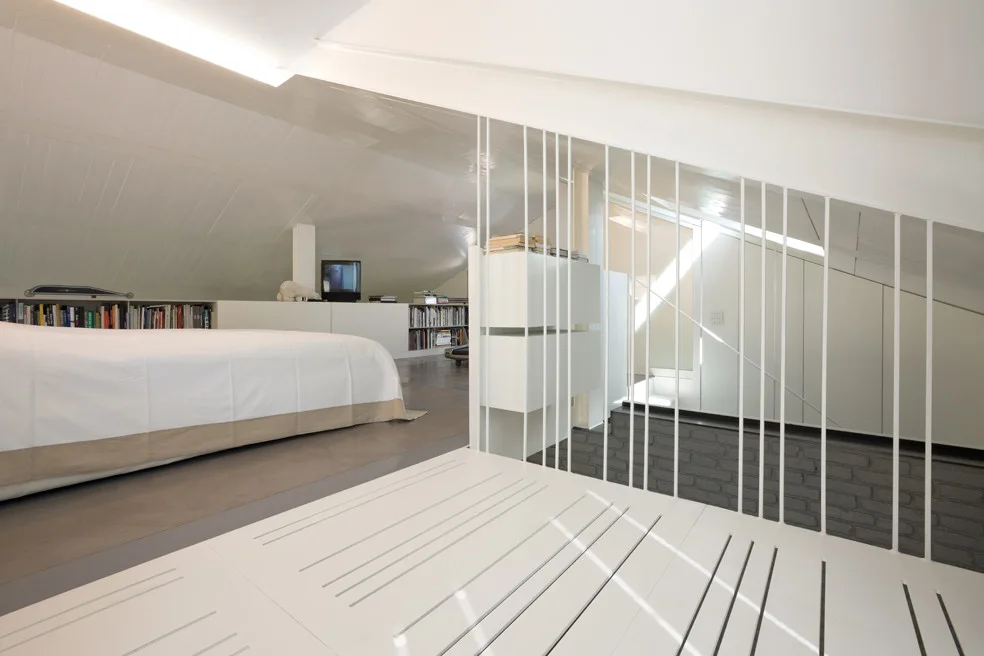 A minimalist attic bedroom features a low bed, built-in bookshelves beneath a sloped ceiling, and a vertical white metal spindle railing.